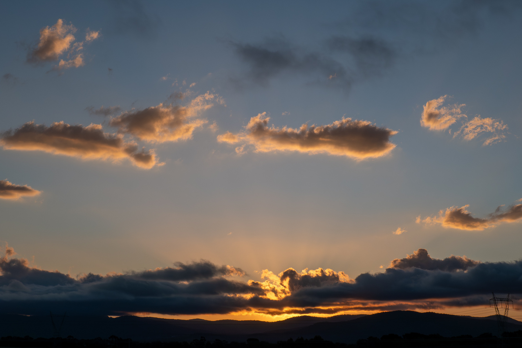 Sunset. There is a band of low clouds just over the mountains on the horizon with a yellow glow under a blue sky, with some small clouds floating past.