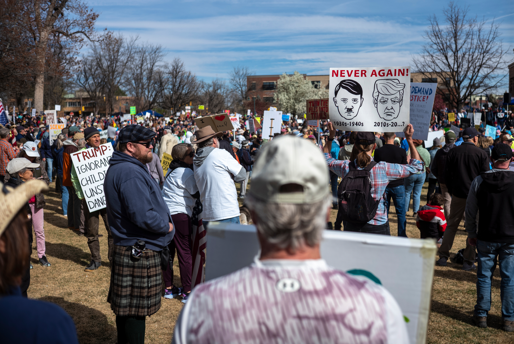 photo of an anti-trump rally with many people in the scene; focus of the image is a person holding a poster that has both hitler and trump drawn on it, with the text 'never again'