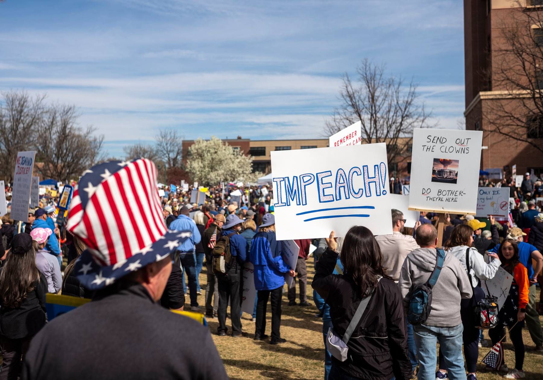 photo of an anti-trump rally with many people in the scene; focus of the image is a person holding a poster that says IMPEACH; person is standing next to another person with a poster that says 'send out the clowns'