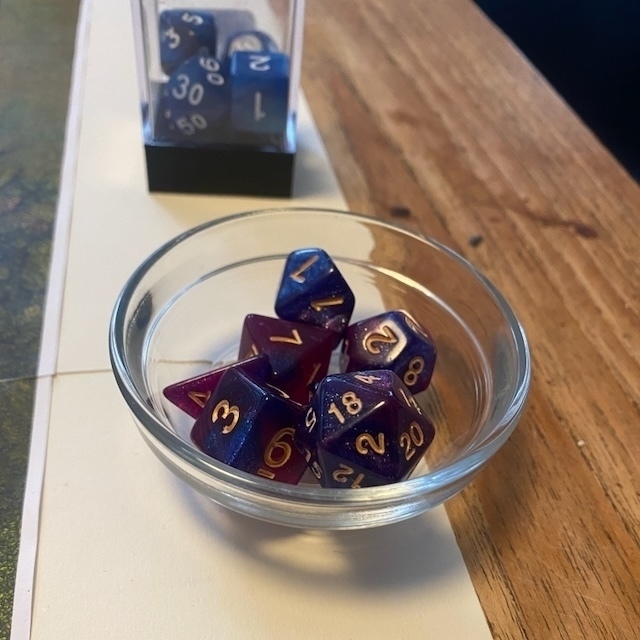 A glass bowl containing colorful polyhedral dice sits on a wooden table, with a clear box holding additional dice in the background.