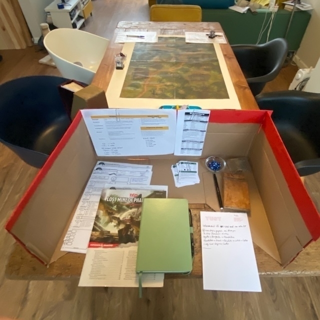 A tabletop setup for a Dungeons & Dragons game with a screen, game materials, and a map laid out on a wooden table.