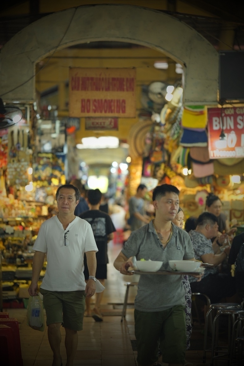 Photograph form the inside of Saigon's most famous markets, where you can buy lots of different crafts, food (and Chinese imports). Sort of colonial architecture, yellowish, and there's a guy running around with two phở. No smoking sign on top