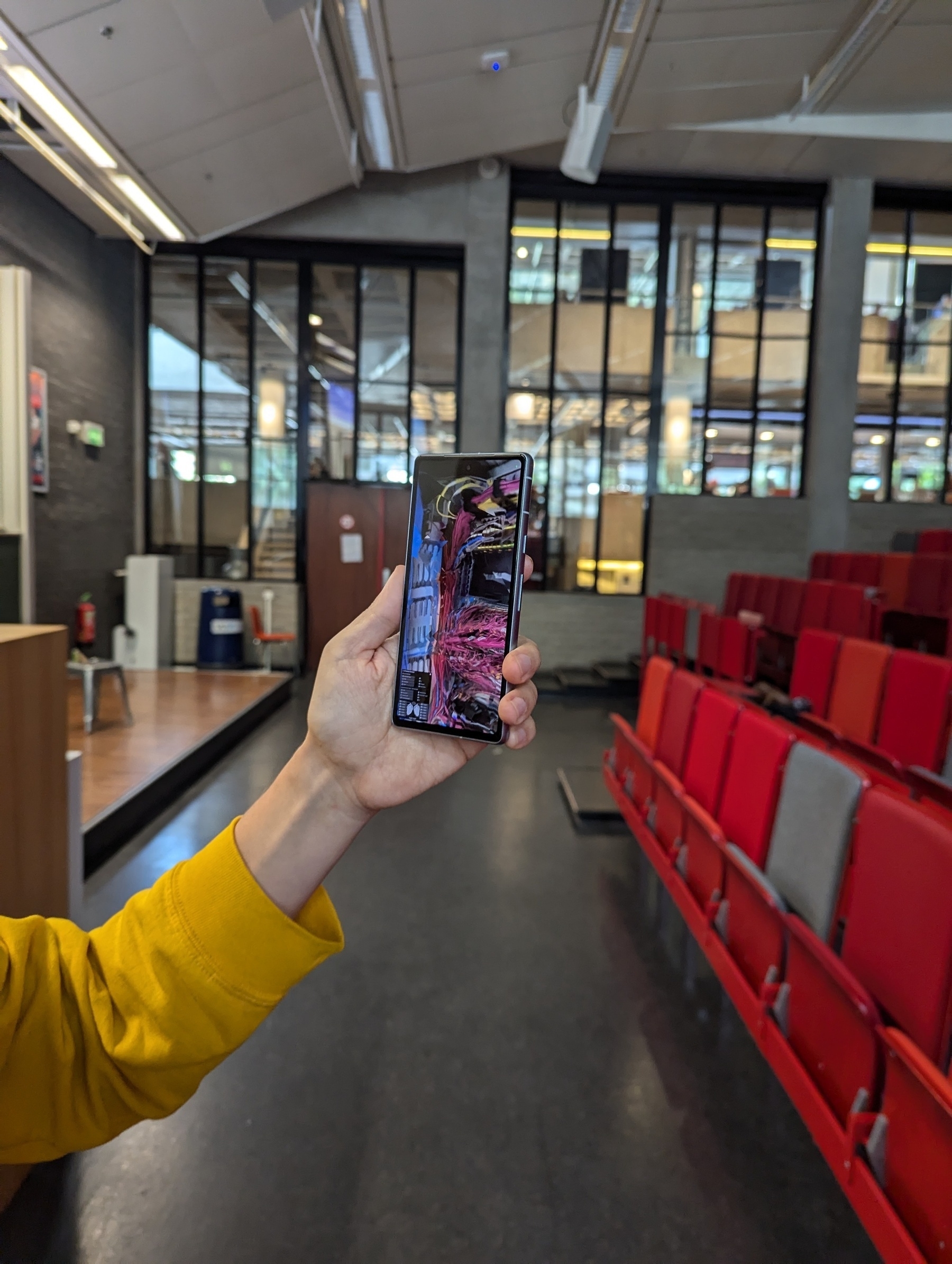 A person in a lecture hall, holding up an artificial reality app that puts a data centre into the cathedral. You see this on the phone. Chalk board area to the left, audience with red seats to the right. And there are hidden Easter eggs in the app 