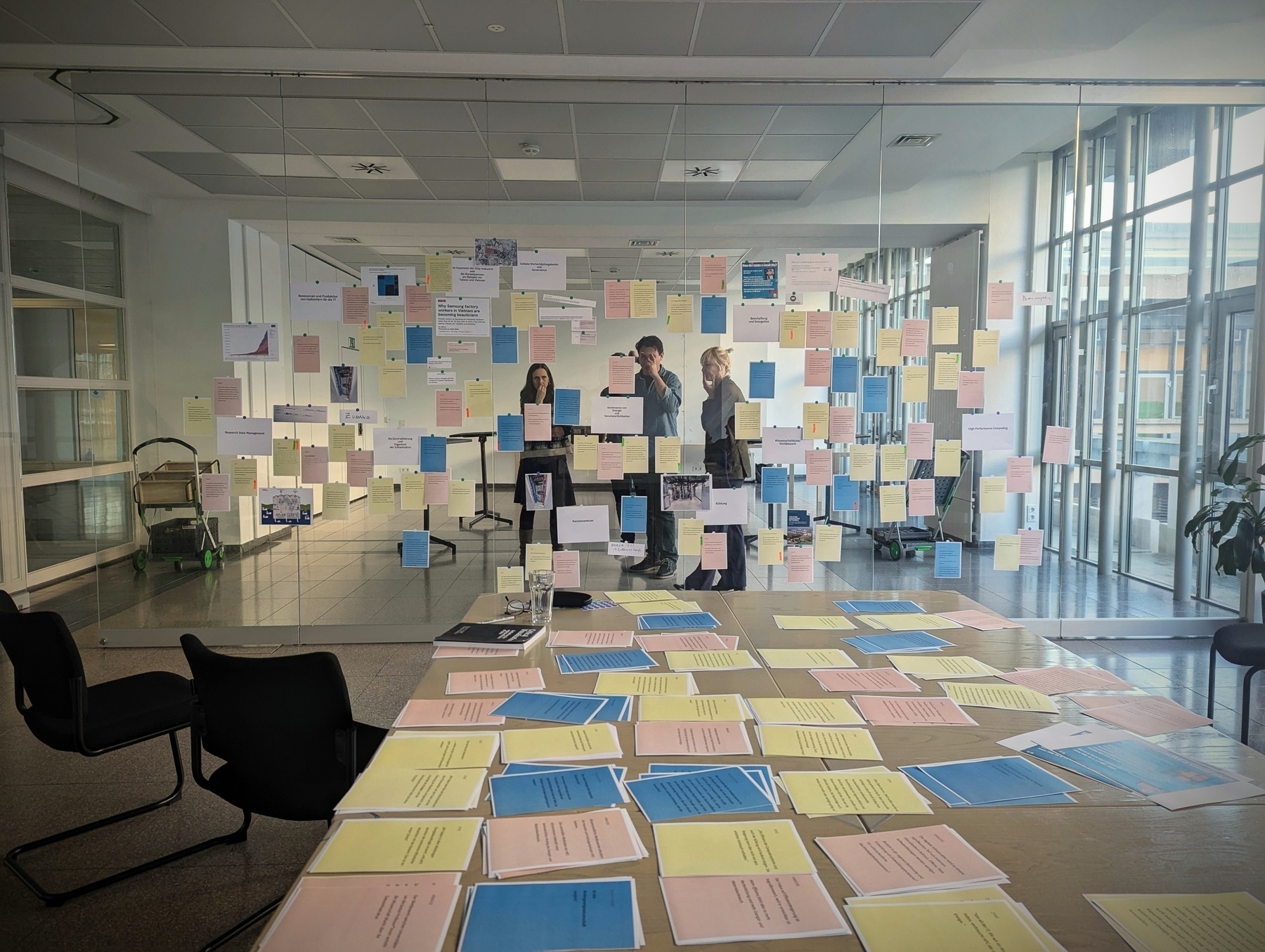 A room with a large table lies in front of you, various A4 pages in yellow, blue and red filling the table. Chaos and structure at once. Behind the table is a glass wall separating the big room, with many notes sticking on the glass front. This is our map, annotated during the event by the actors. Transparent mapping. Behind the glass wall, a few people stand and look at the notes from behind, reflecting on the implicit structure. Far behind, there is the actual wall of the large room, with coffee etc. Waiting to be digested. 