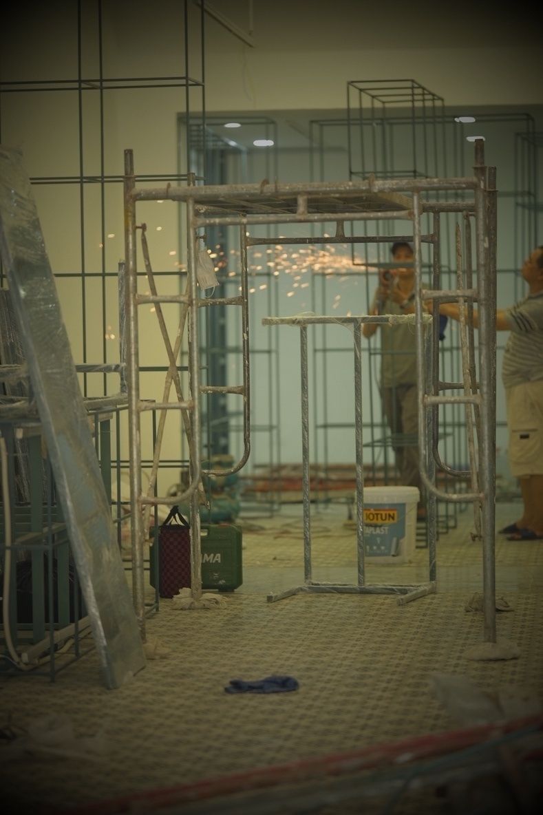 Photograph into a newly built shop. Lots of pols. Construction is going on, a worker cuts metal, spraying sparks. Another worker watched. Nice flooring.