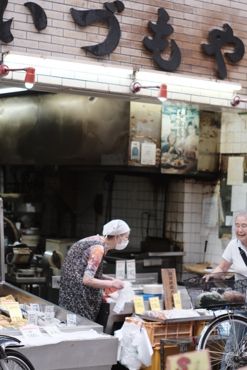 Two old women enjoying a chat while the left one packs a serving of tofu. She buckles a bit, and there are lots of tiny price signs around for the various tofu things to get, and the wall of the store is a bit roasted from the cooking that is going on all the time 