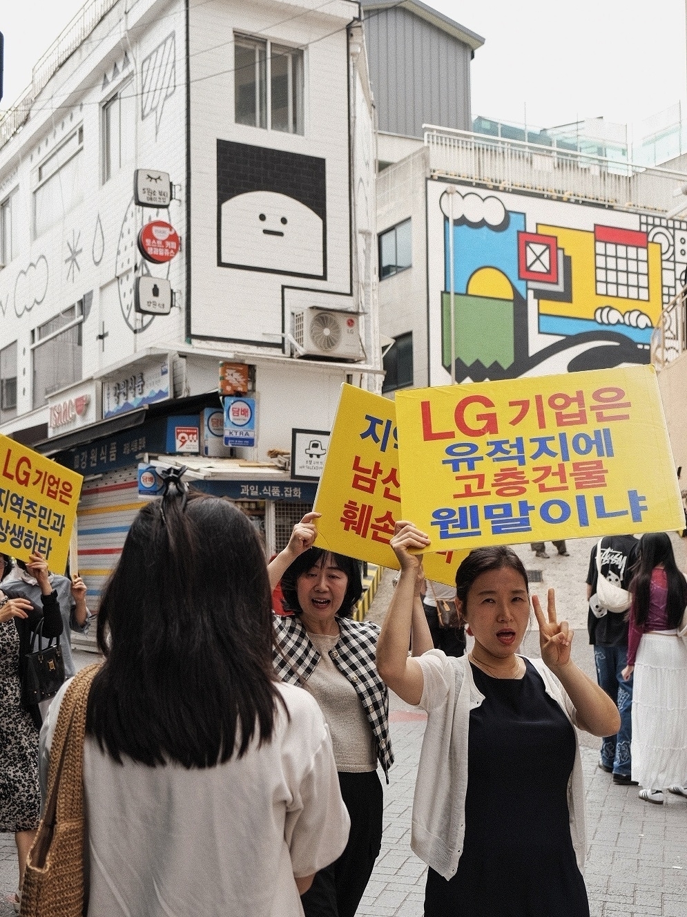 Protesters holding signs against LG. Red and blue Korean script on yellow signs, local buildings and hills in the background 
