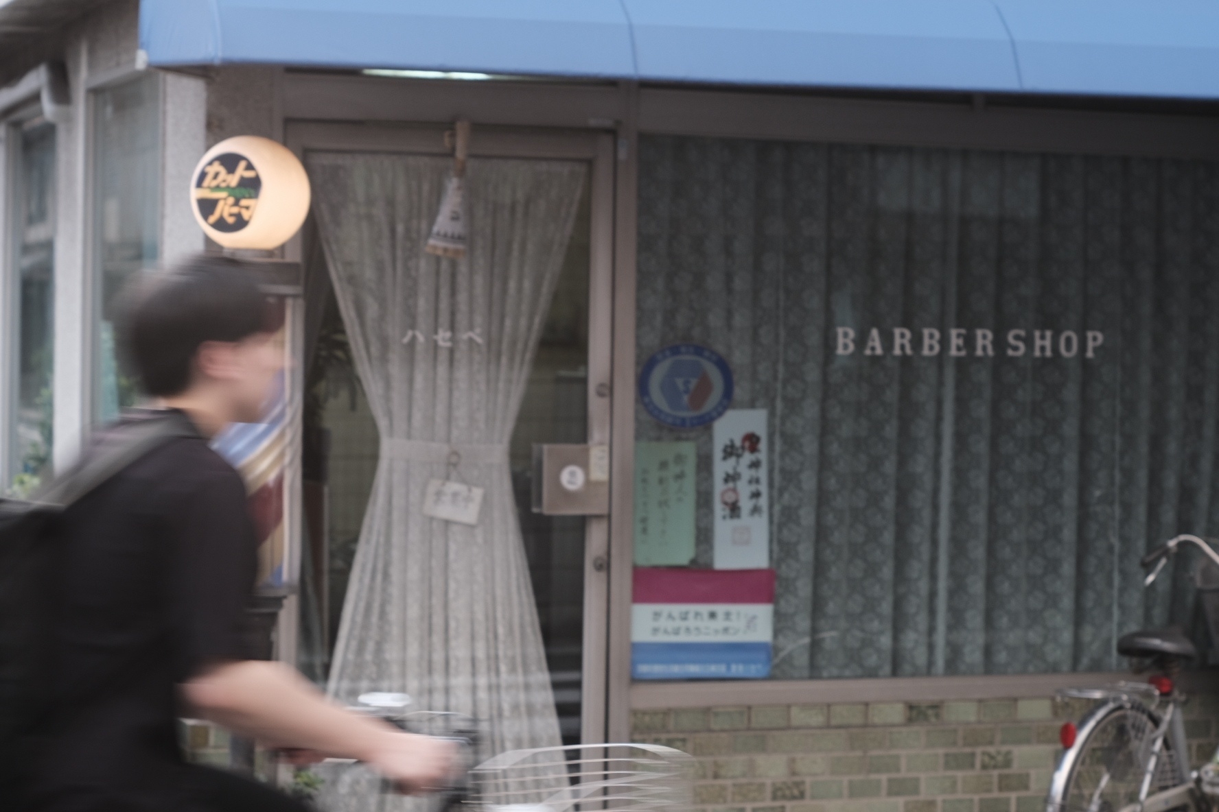 A sign of a barber shop with the iconic barber colours. Grey curtains next to a grey brick wall. And then there's a cyclist just riding into the frame form the left 