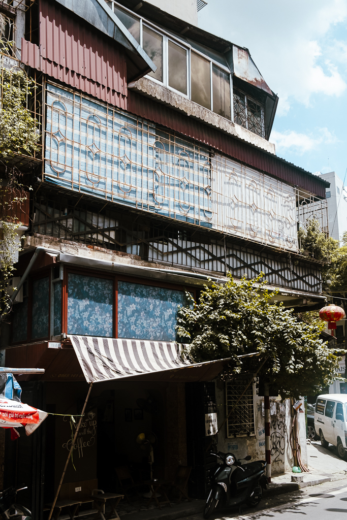 Vietnamese house and coffee shop shot from the side with a blue sky in the black. The house front has many layers of different colourful fronts and corrugated iron in between. A few windows on top. Lots of green flowers overflowing. An inviting sight