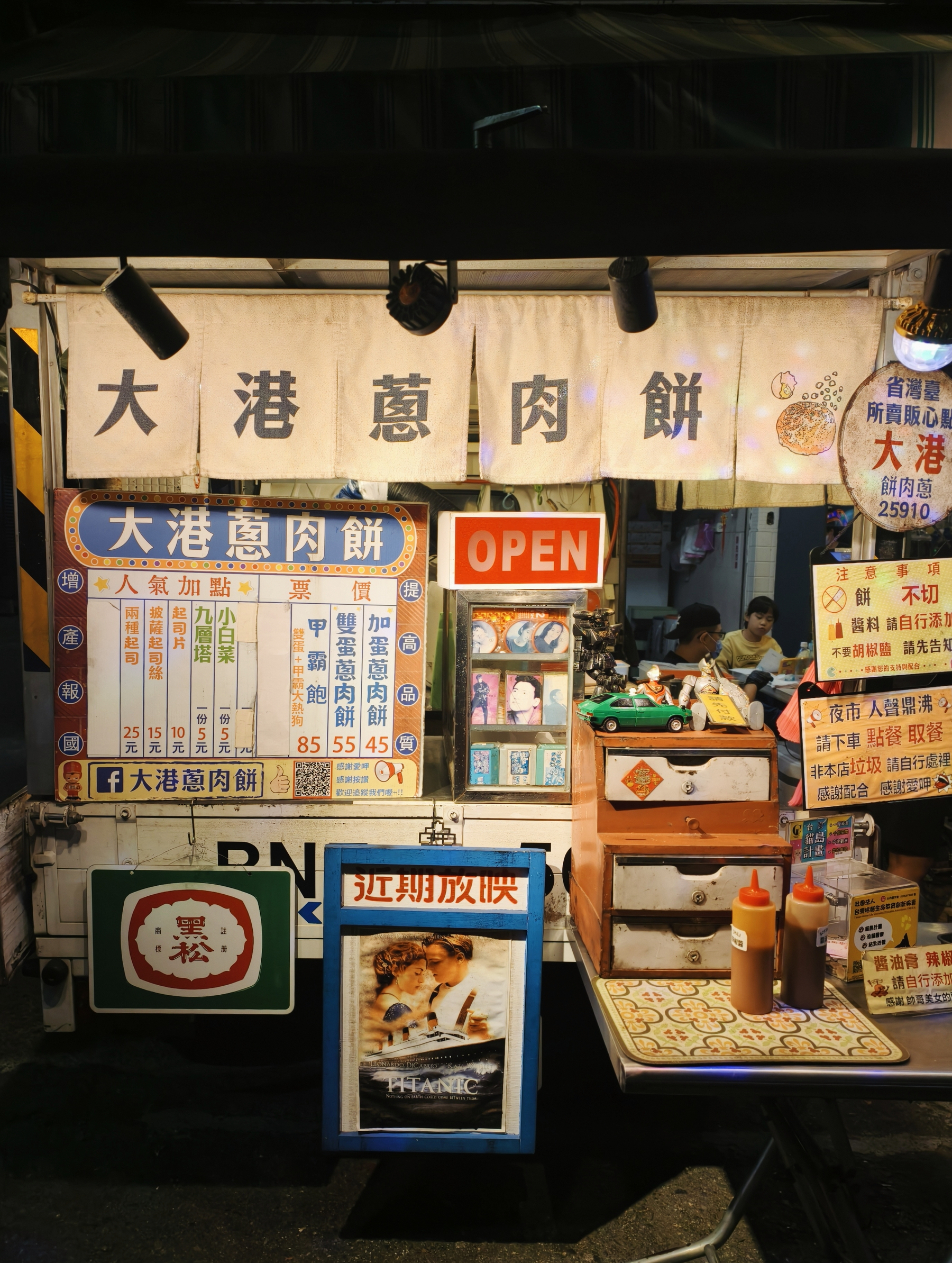A night market stand selling meat pie with colourful yet tasteful advertising. Also, a poster of Titanic in the front 