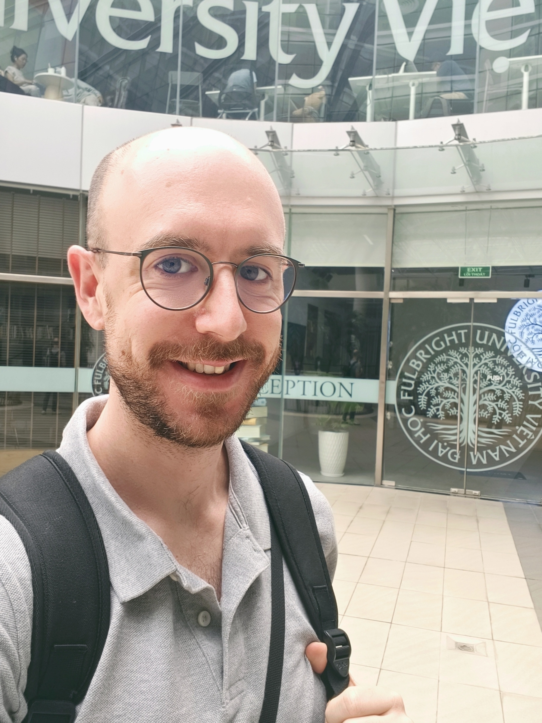 Picture of a bald men with a beard, me, smiling in front of the library of Fulbright Uni