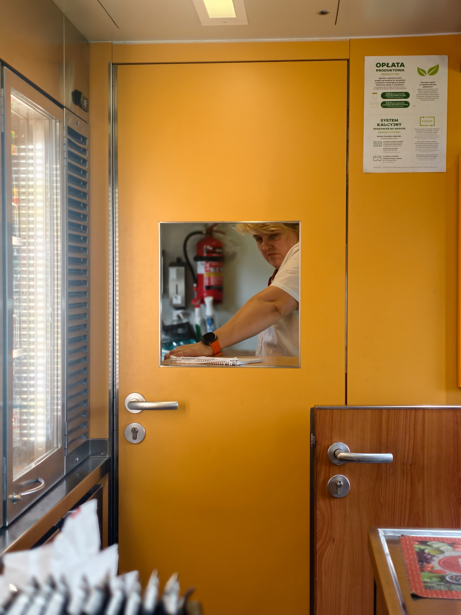 The actual kitchen on board where food is prepared; a photograph of the woman looking through a small window in the door, yellow interior