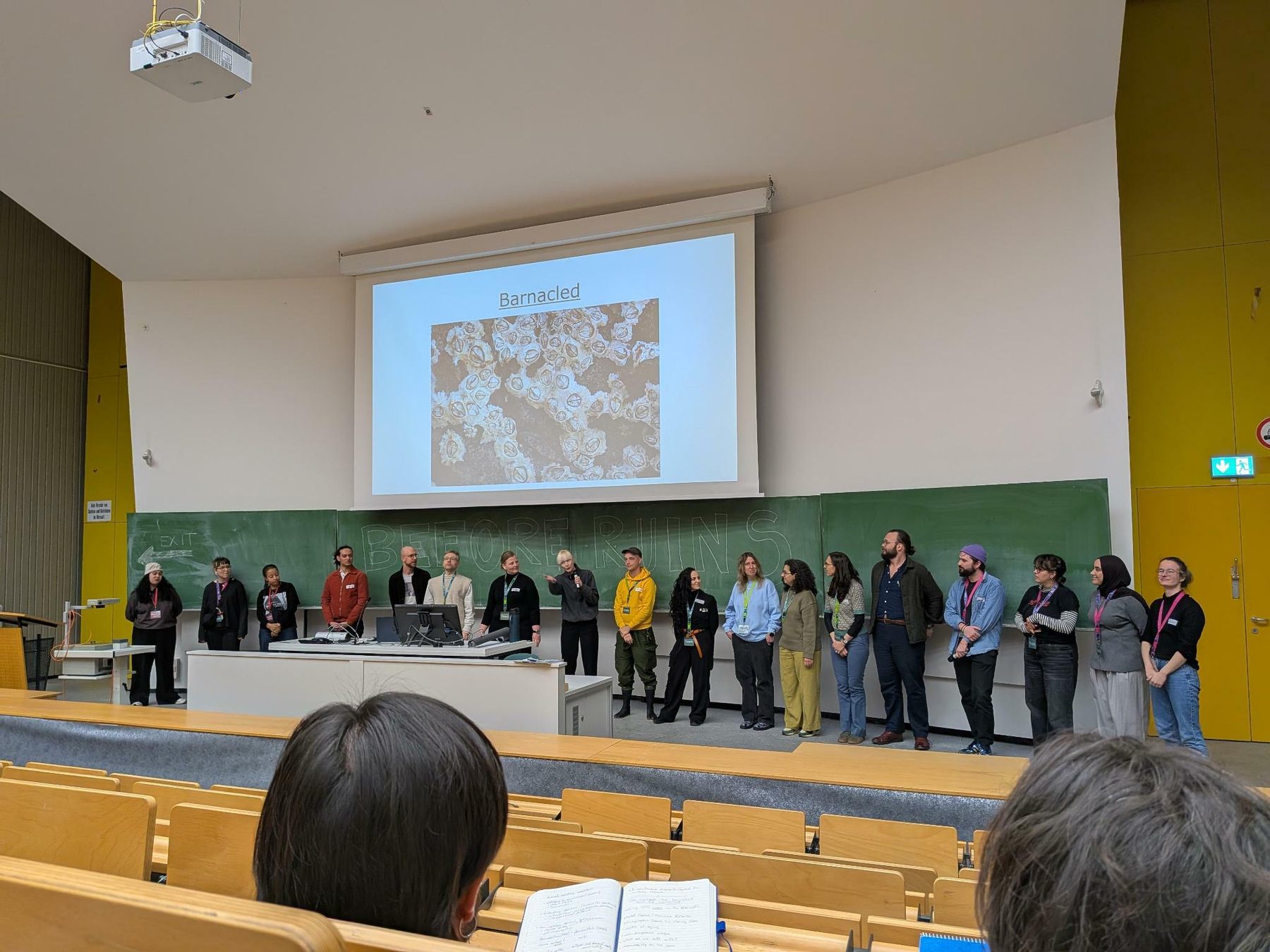 Our team of organizers and student assistants on stage waving a good bye. 18 people in a lecture hall, the photo is shot from the seating area.