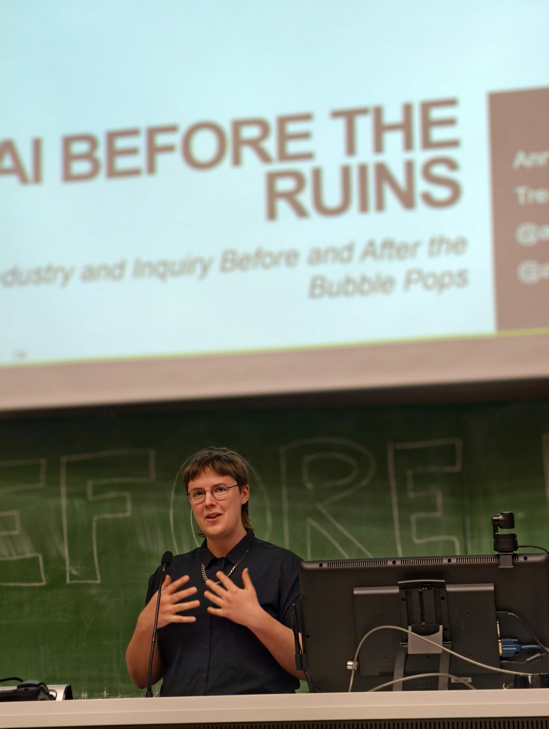 Keynote photo of Anne. A woman presenting standing next to a computer screen, the slides and a green chalkboard behind