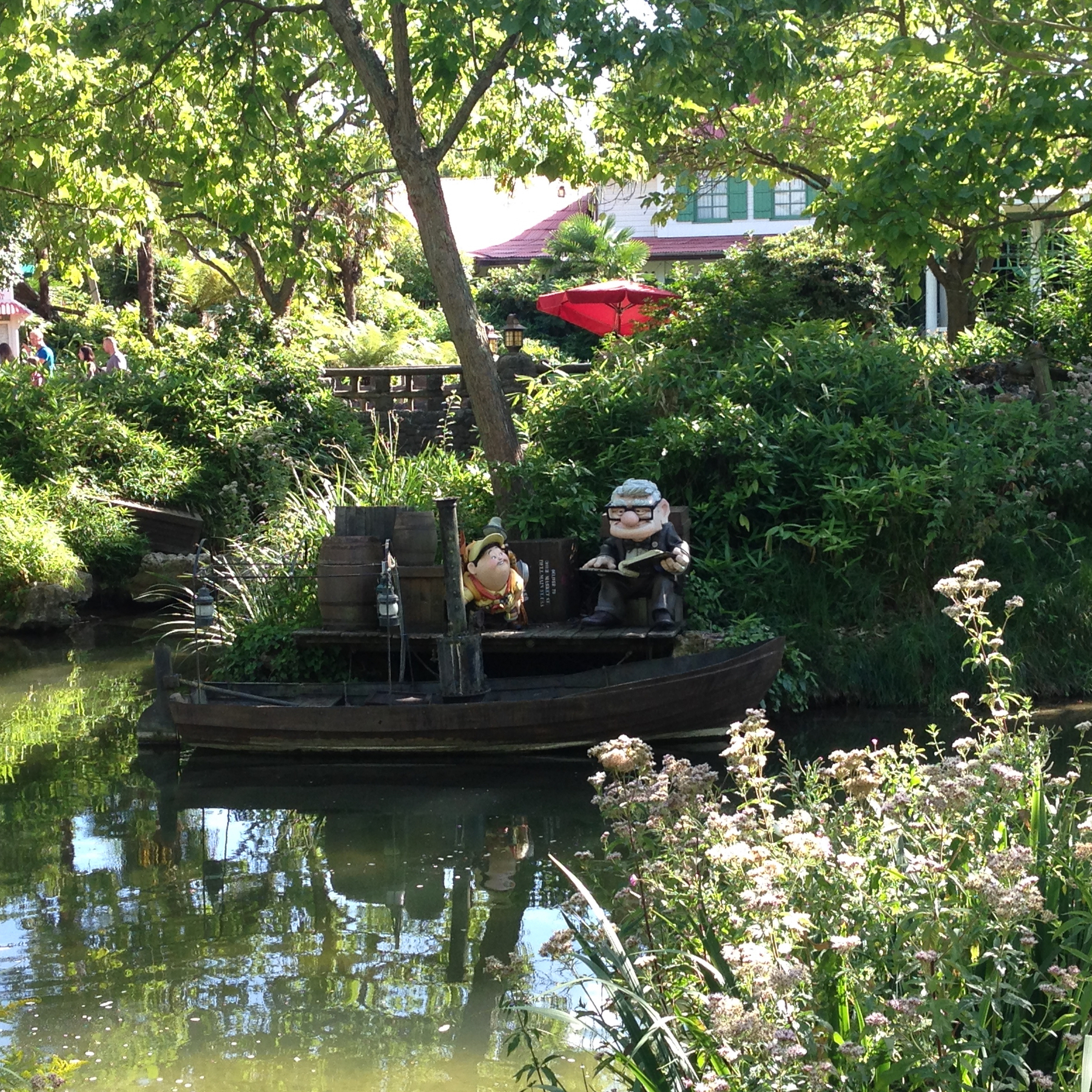 Two cartoonish statues of elderly characters are seated on a small boat surrounded by greenery and a calm pond.
