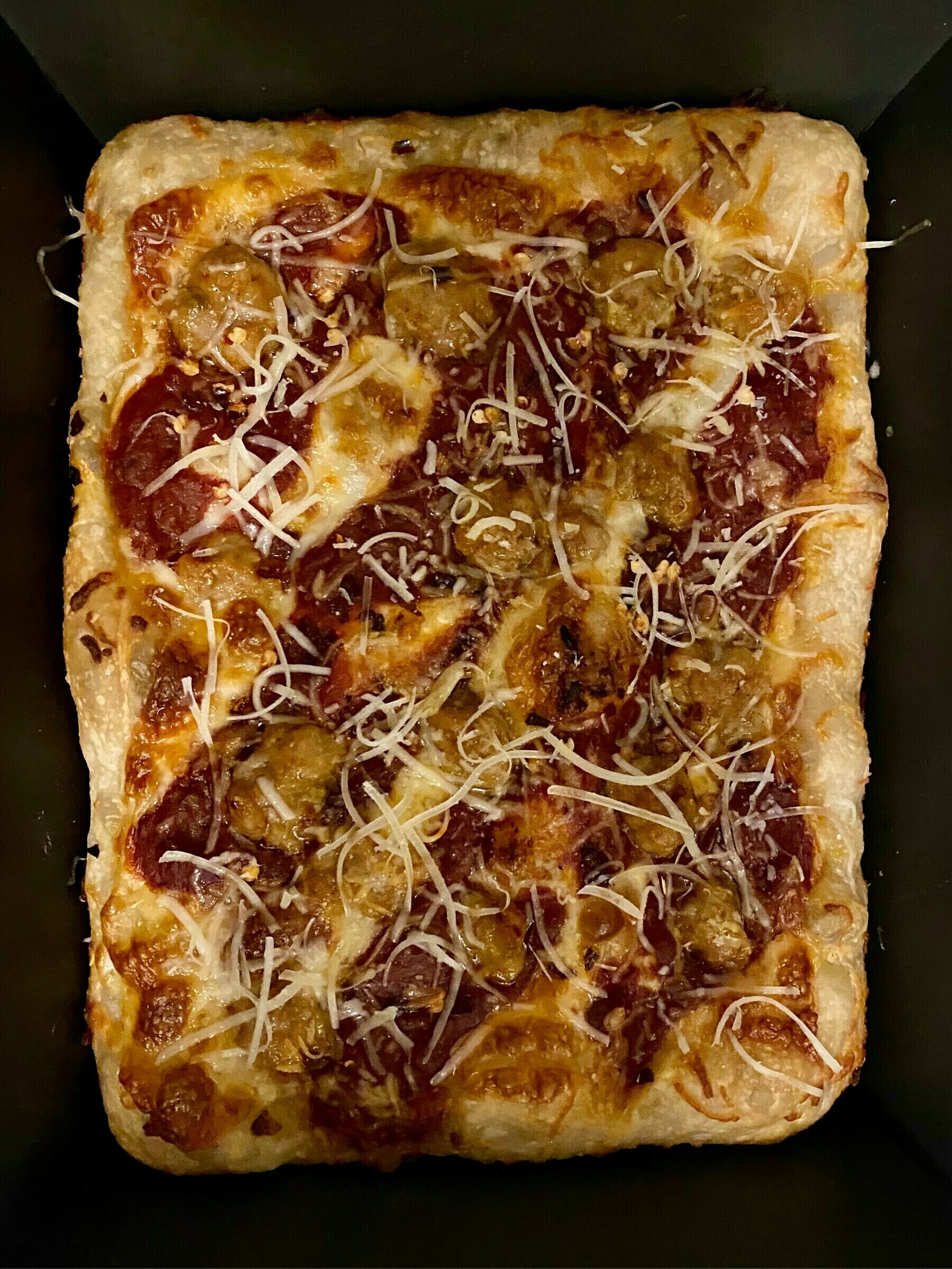 Top down view of an 8 by 10 inch pan pizza cooling in the pan. It has a sliced mozzarella base, stripes of tomato sauce, bits of sausage and shredded parmasean cheese on it.