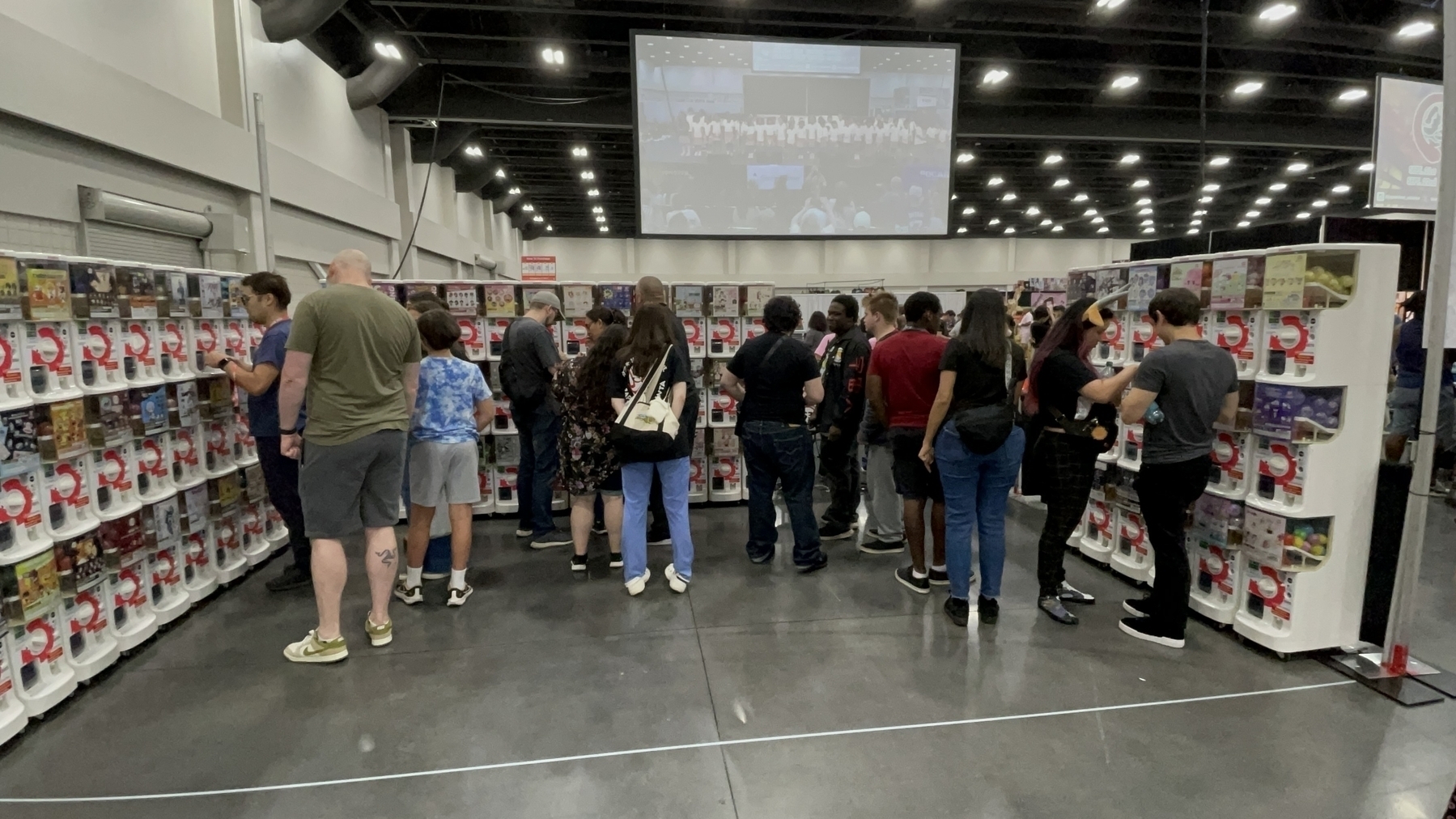 A group of people are browsing shelves filled with “goshapon” capsule machines in a large indoor space.