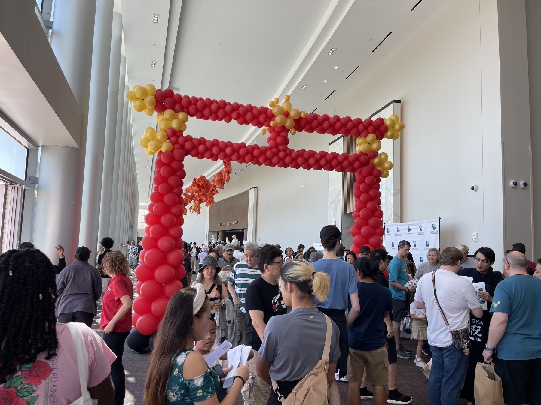 A crowd of people gathers near a large Japanese “Torii”archway made of red and yellow balloons inside a spacious, brightly lit room.