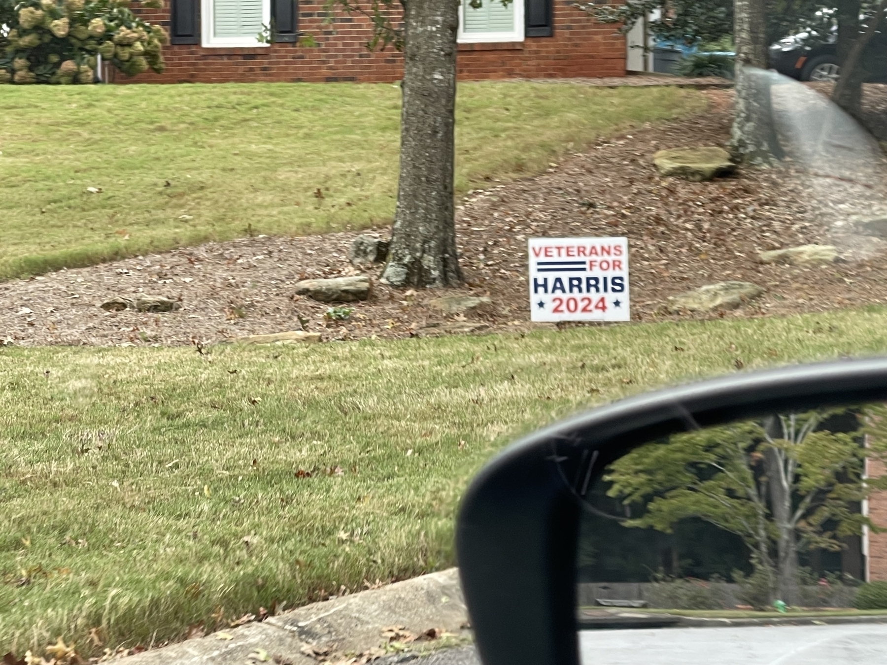 A campaign sign reading "Veterans for Harris 2024" is placed in a grassy area near trees in a suburban home's yard.