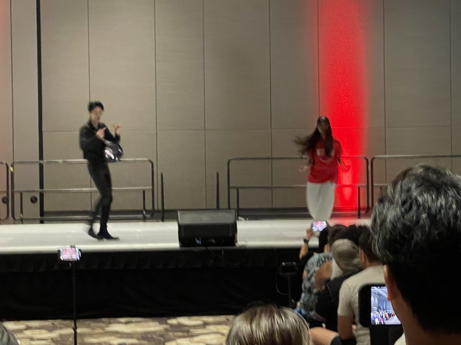 Musicians play traditional Japanese instruments on stage while a crowd watches, and two speakers stand nearby with microphones.