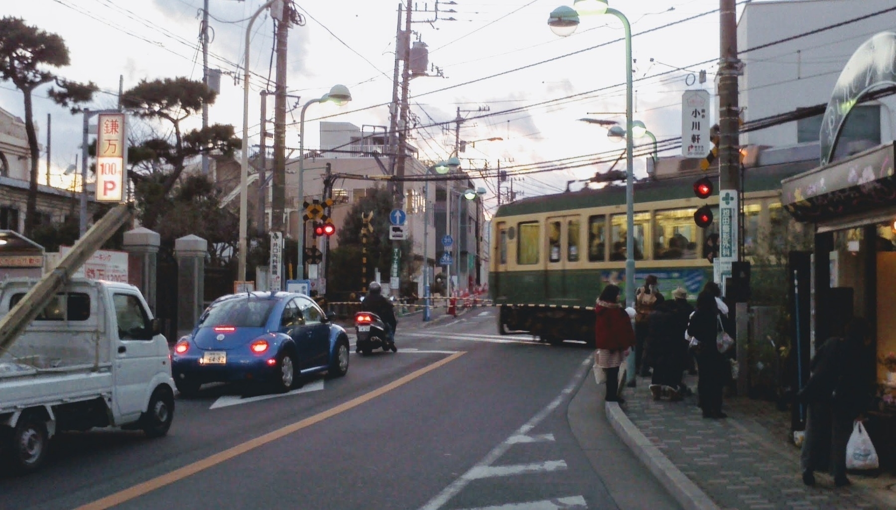 A Japanese street scene features a train crossing the road, several vehicles waiting, and pedestrians walking nearby.