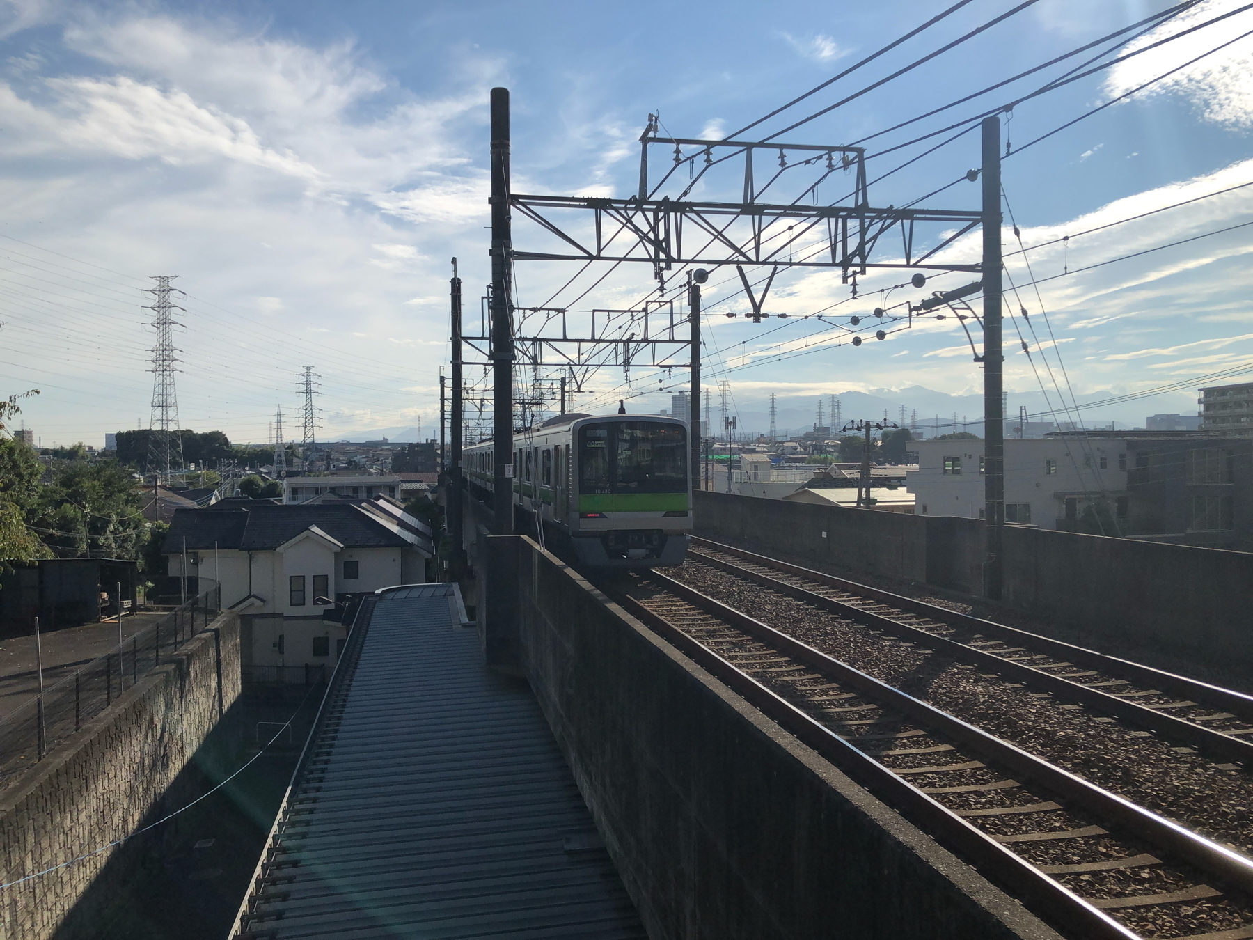 A train travels on elevated tracks under a partially cloudy sky with power lines and buildings in the distance.