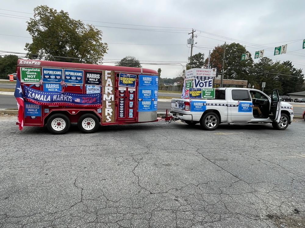 Auto-generated description: A vehicle and trailer are covered with numerous political campaign signs in a parking lot area.