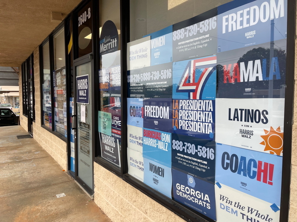 description: A storefront window is covered with a variety of Harris/Walz campaign posters and political advertisements.