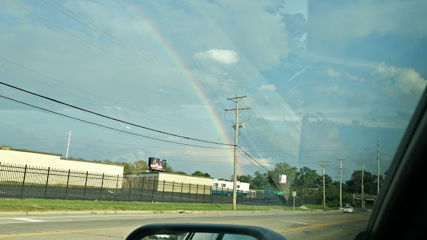 Rainbow in a blue sky as seen over an industrial park in St Louis County, Missouri, USA. The photo was taken from the driver side window of a car and part of the mirror is seen.