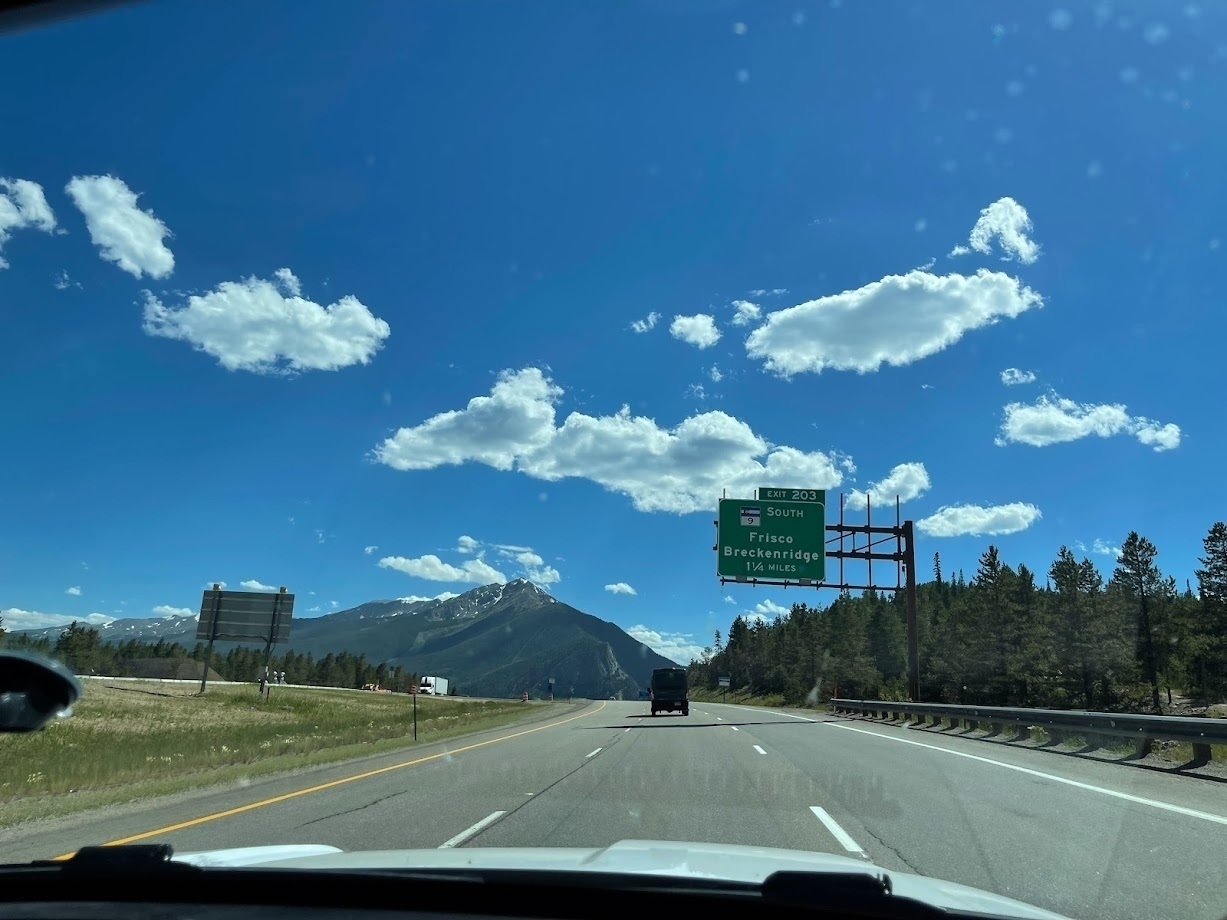 View from a car driving on a highway with clear blue skies and scattered clouds above, featuring a green road sign for Exit 203 to Frisco and Breckenridge with mountains in the distance.