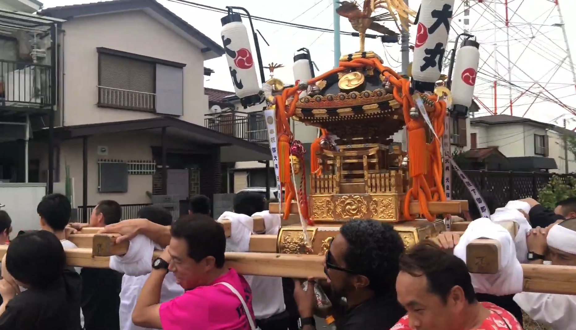 The image depicts a traditional Japanese festival scene where participants are carrying a mikoshi (portable shrine). The mikoshi is elaborately decorated in gold with white lanterns and red accents. Participants are dressed in traditional festival attire, some wearing headbands, while others are in casual clothes. They are lifting the mikoshi on their shoulders using two long wooden beams. In the background, there are residential buildings and power lines under a cloudy sky.