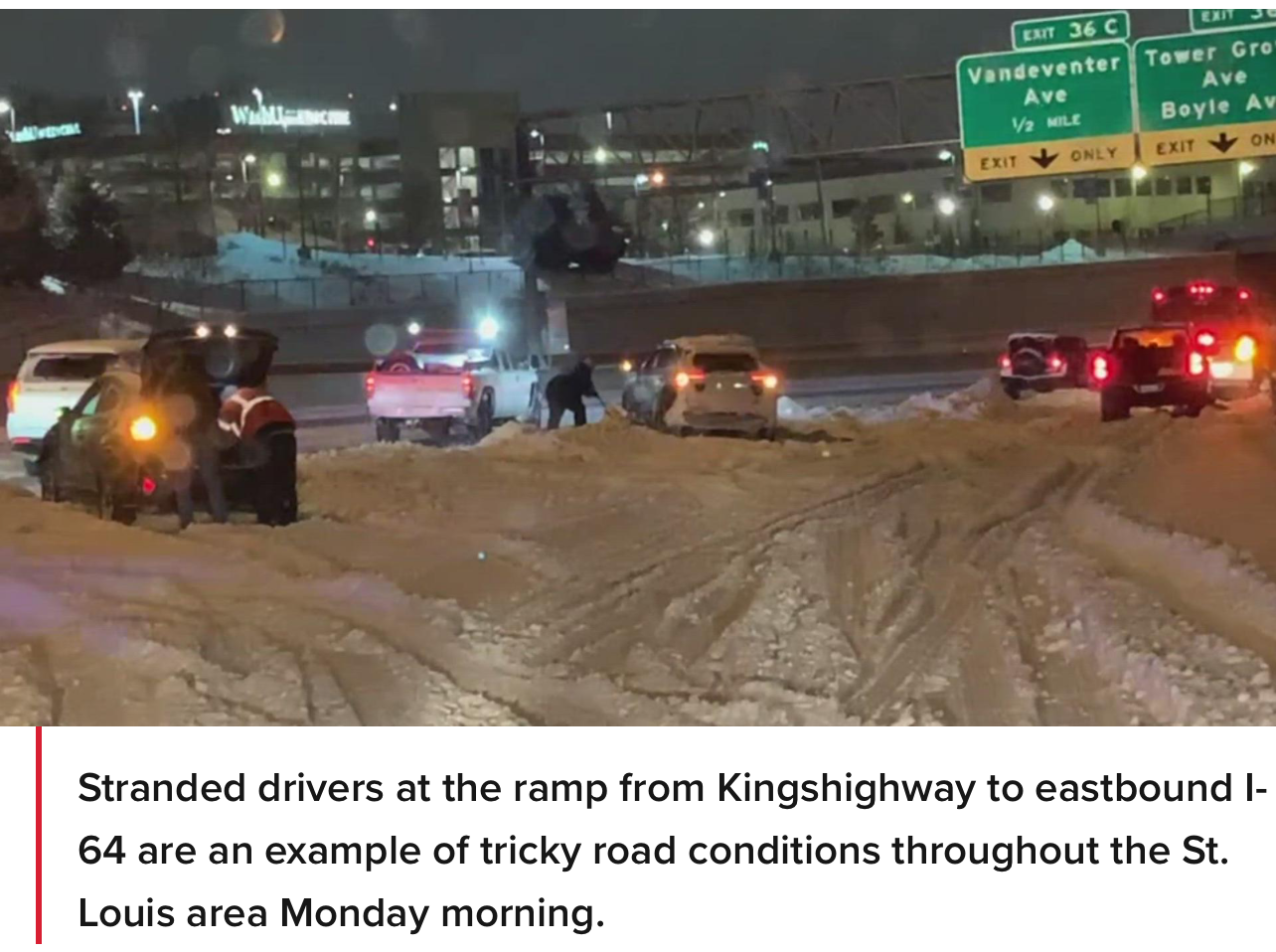 Stranded vehicles and people attempting to push cars are seen on a snow-covered ramp leading to a busy highway at night.