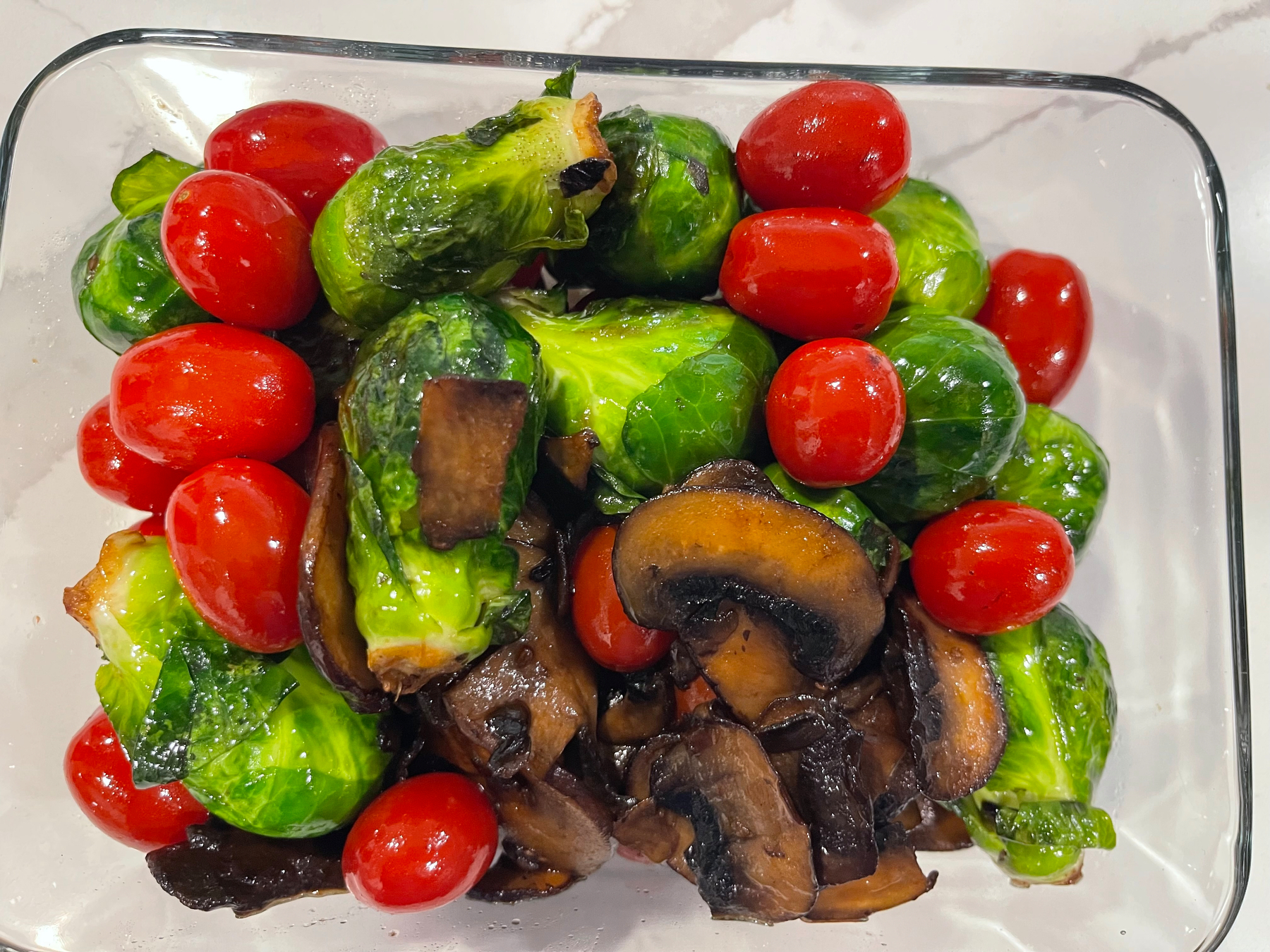 A dish featuring Brussels sprouts, cherry tomatoes, and sautéed mushrooms is arranged in a clear glass dish.