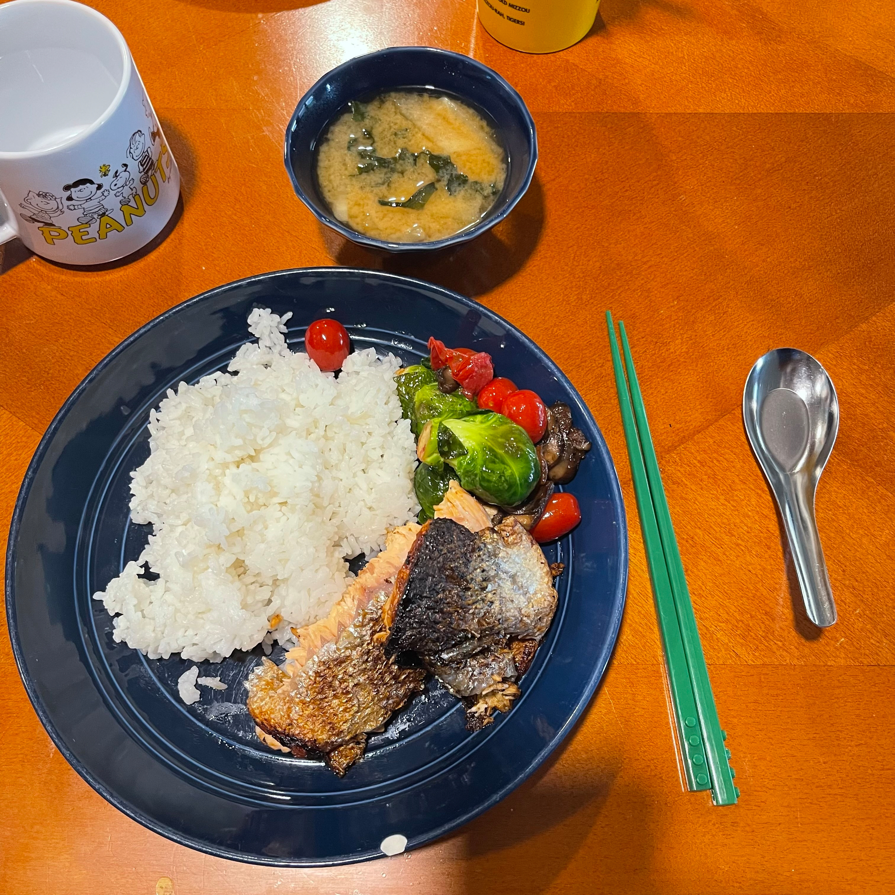 A meal featuring a plate with rice, grilled fish, Brussels sprouts, and tomatoes is served alongside a bowl of miso soup, green chopsticks, and a mug on a wooden table.