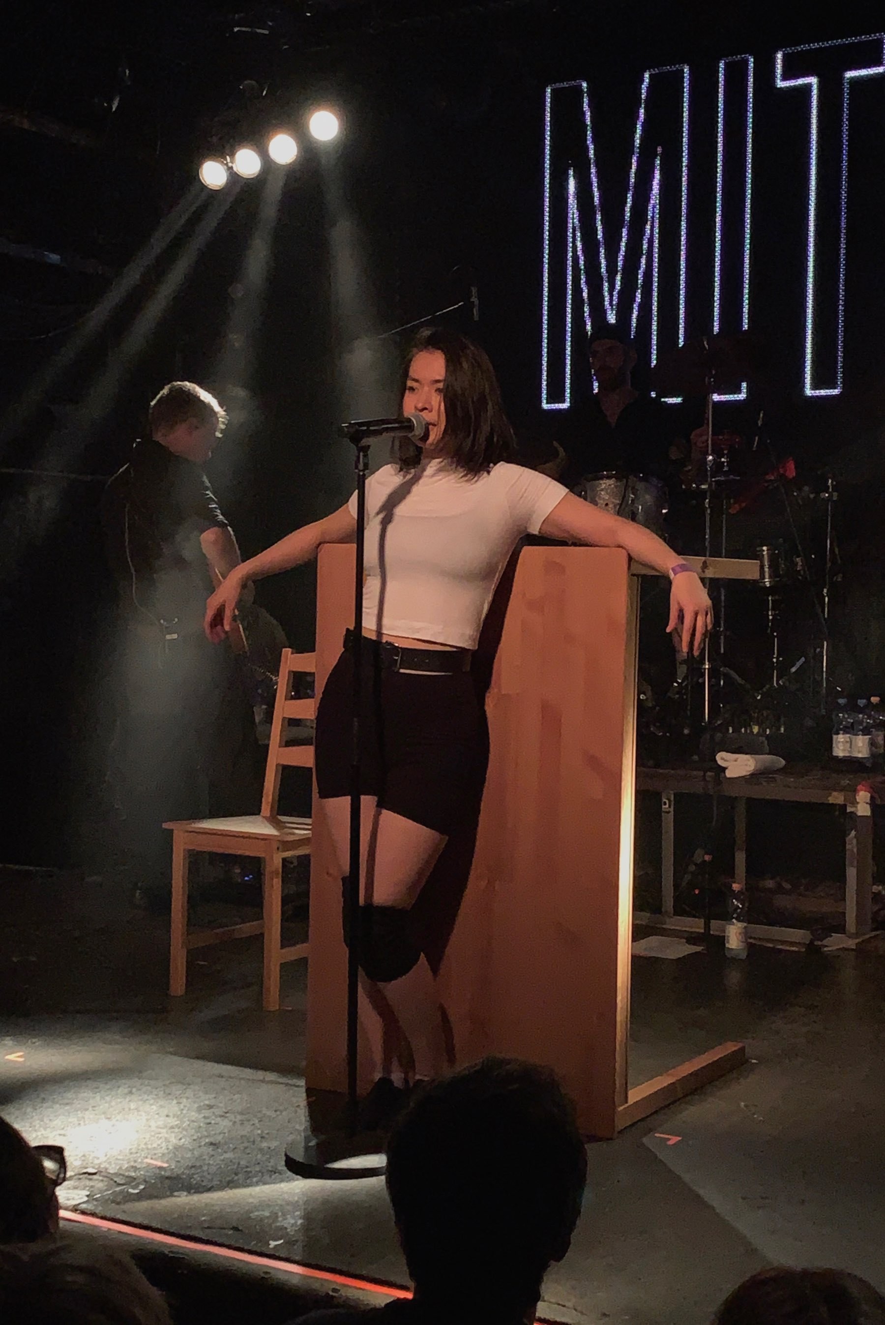 Mitski singing and leaning on a table with part of her band in the background on stage at Flex