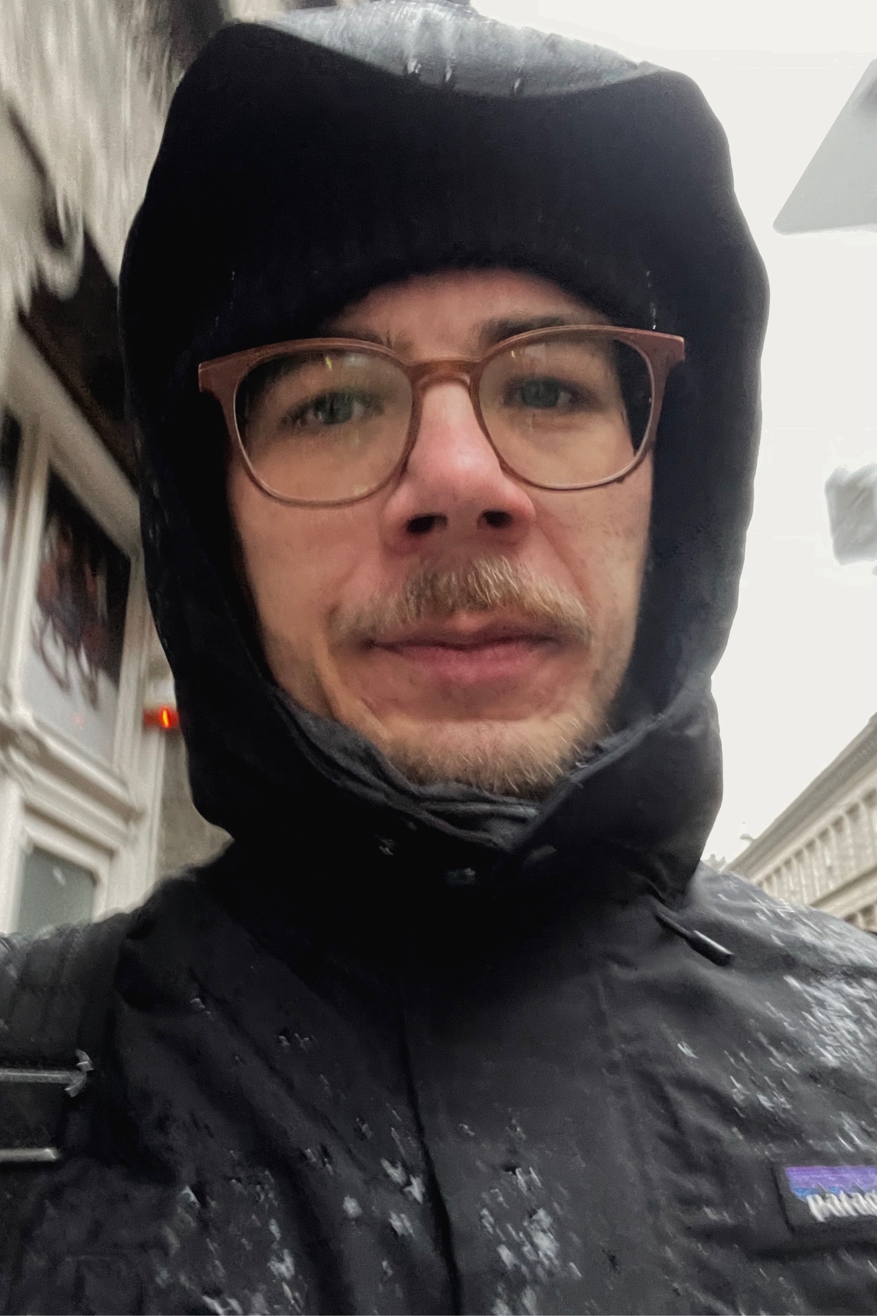Selfie of a man walking in the rain, wearing a black wool hat and a black coat with a hood. there are drops on his glasses and he looks very wet.
