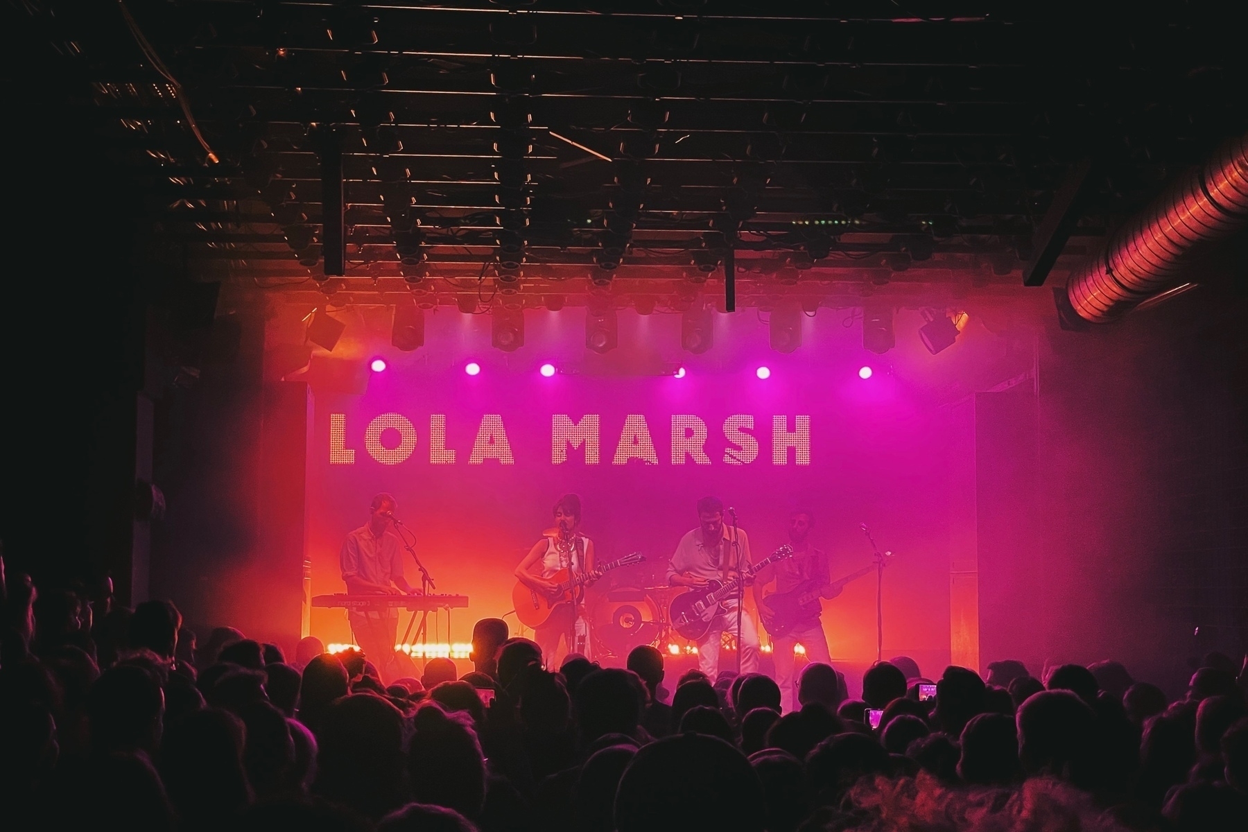 The band Lola Marsh on stage at Flex in Vienna, the singer is in the middle, on the left a keyboard player, guitar and bass player on the right and drums in the back. The light is pink-yellowish, in the back of the stage the band name is written. In the foreground the crowd is visible.