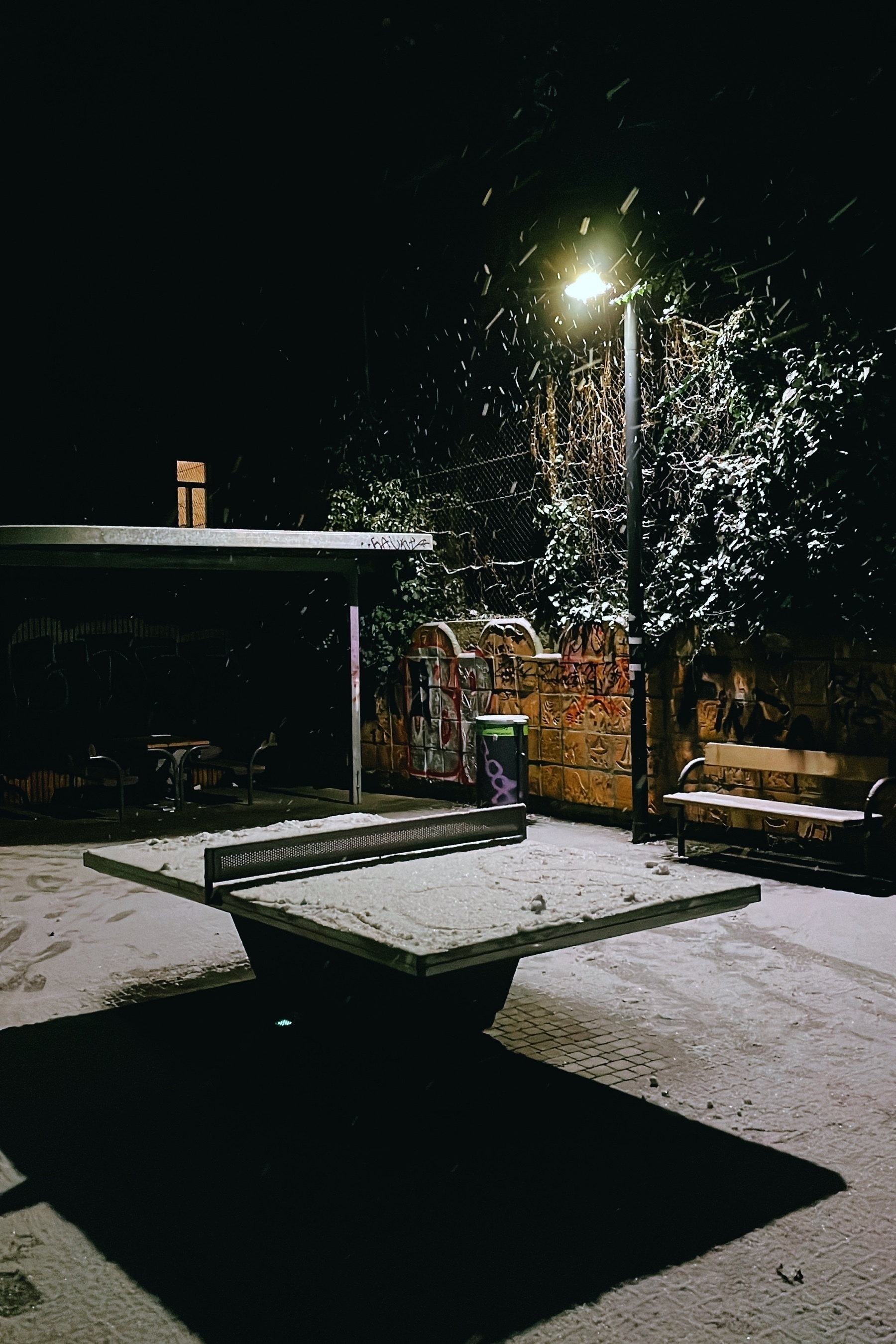 Table tennis table in a park at night, illuminated by a street light, snow is on the ground and table. A brick wall is in the background.
