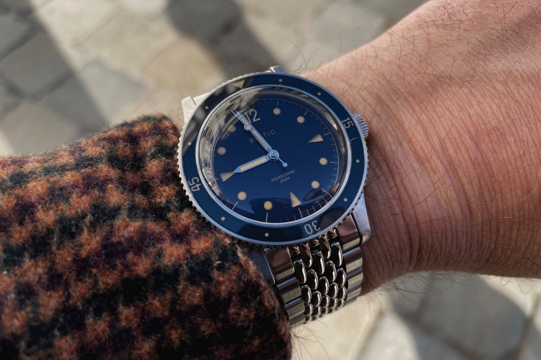Man’s arm with a brown flannel shirt sleeve and a dive watch from Baltic with a steel band and a blue dial.
