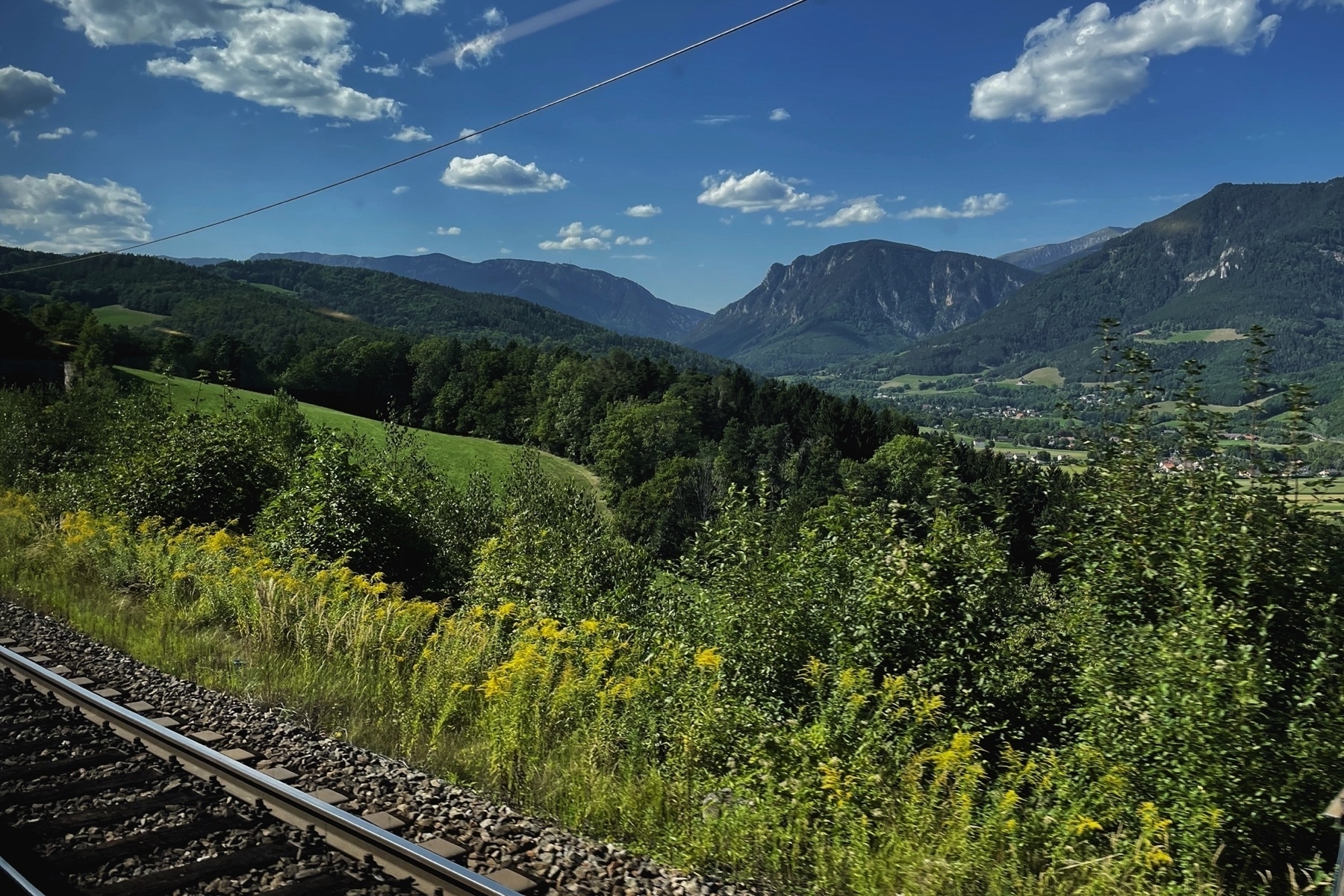 View of a train window, parts of the tracks in the foreground, in the back a valley with hills surrounding it and lots of forests