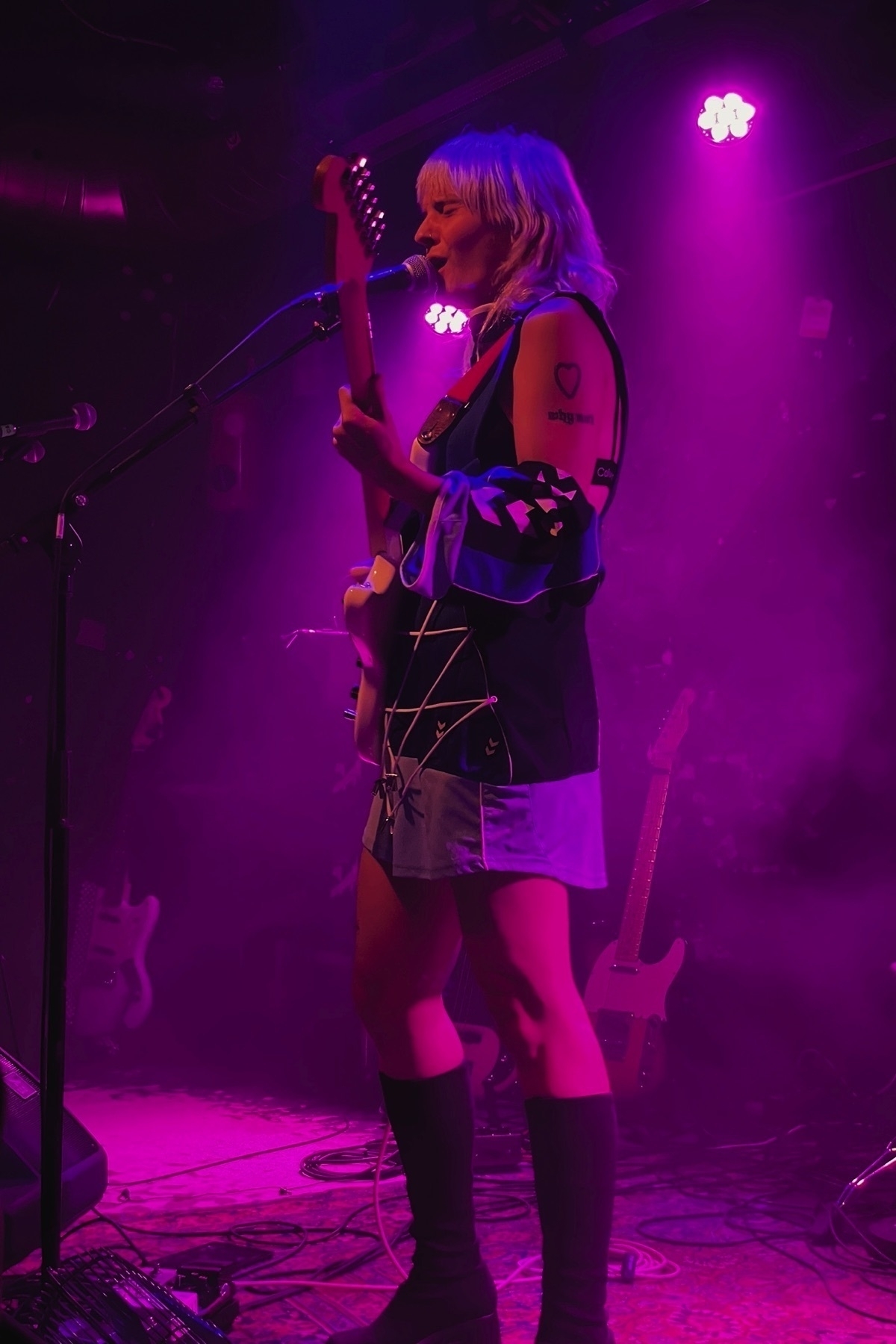 Woman playing guitar and singing into a microphone while standing on stage, the lighting is violet.