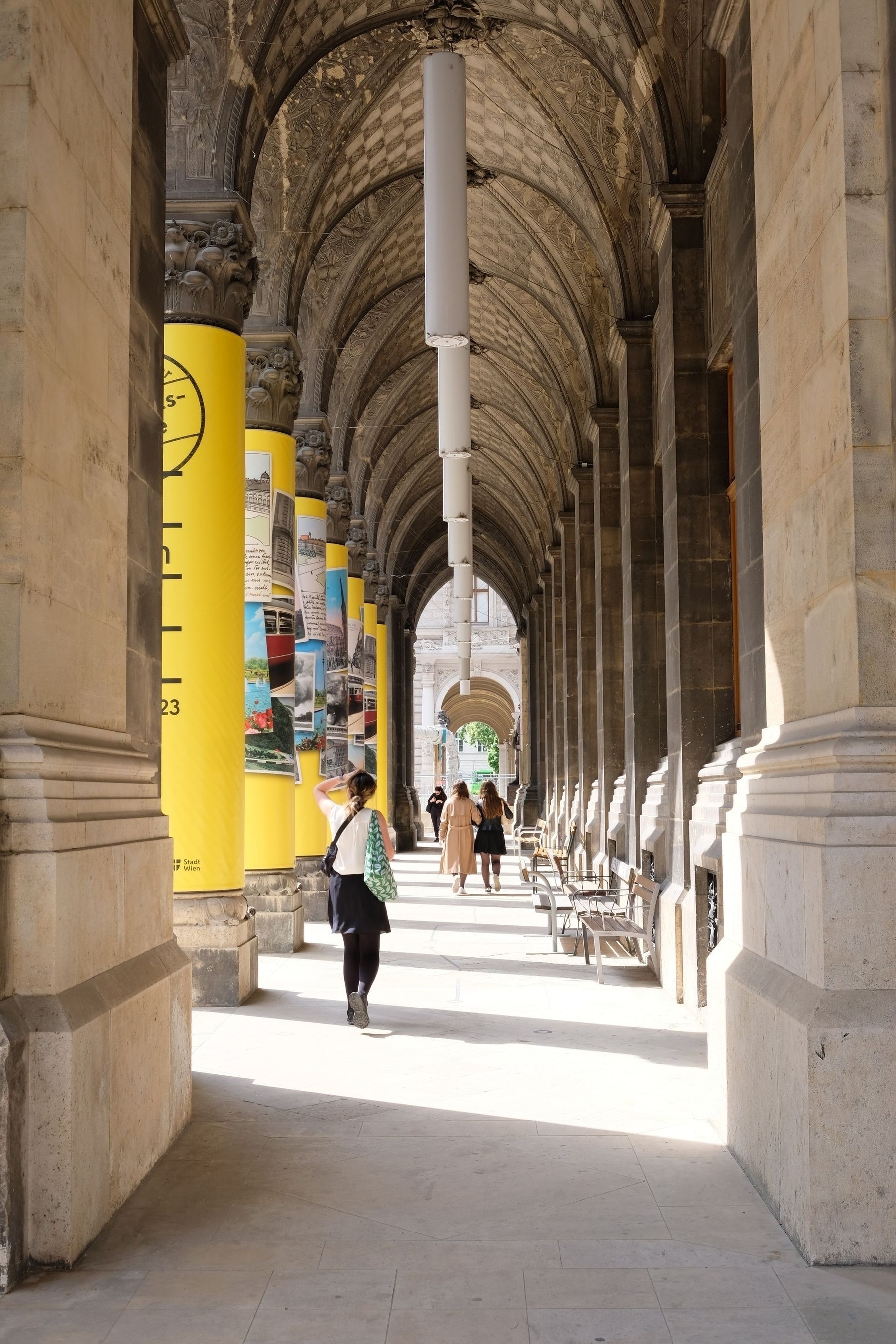 Stone arcade, on the left are columns covered in yellow posters, the building is rather old, a couple of people are walking through the arcade with their back to the camera; the sun is shinning