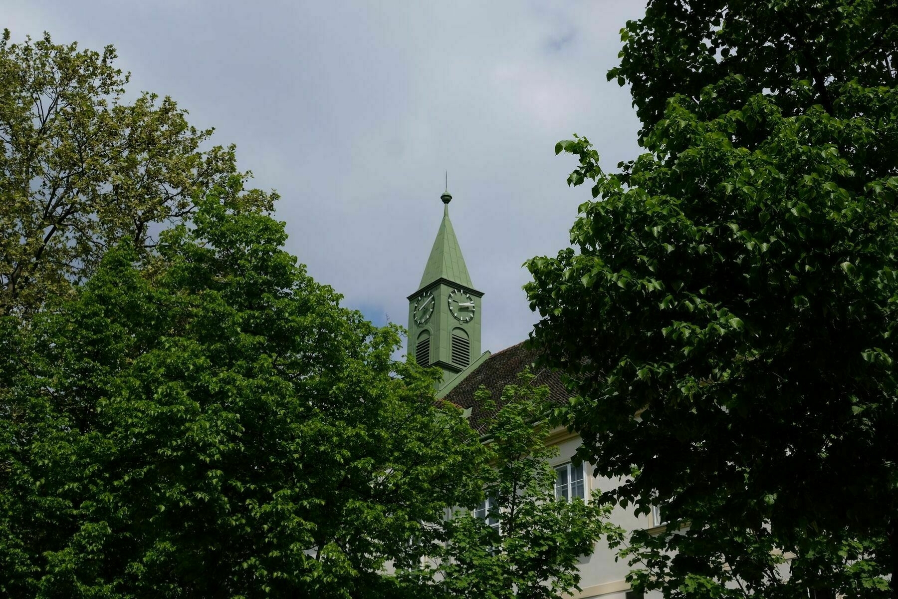 Building with a green turret with a clock on each side, most of the building is hidden by trees, but the turret is in clear view in the center