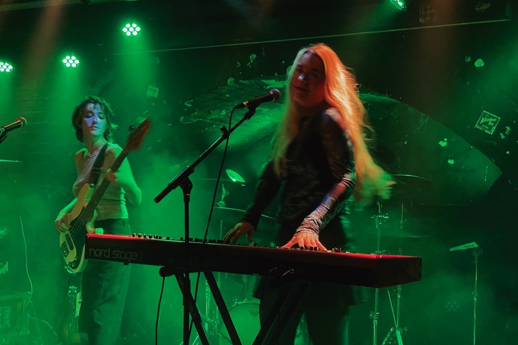 Woman playing piano and in the middle of a turning motion looking into the camera; on the left another woman playing bass guitar, the light is green