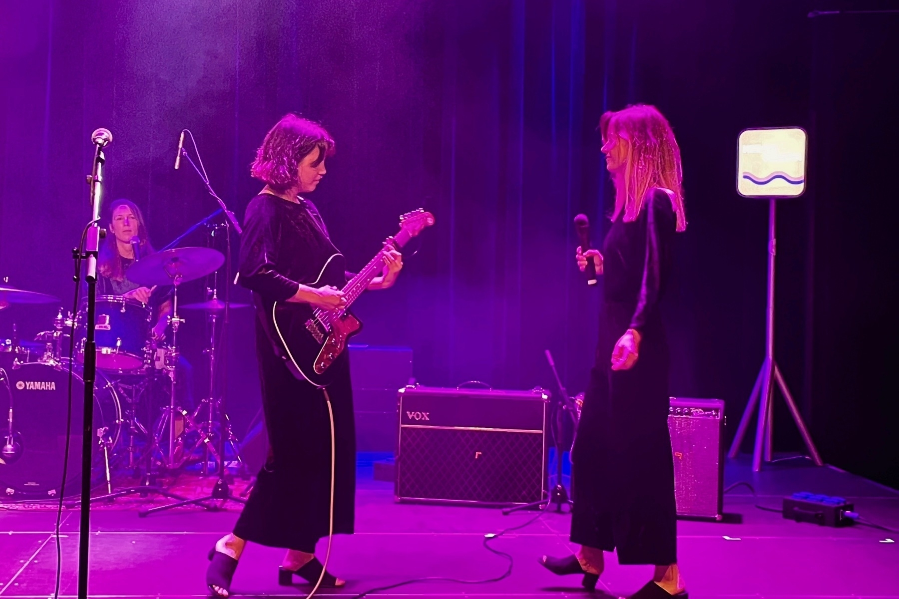 Guitarist and singer facing each other, both wearing the same velveteen onesie. In the background the drummer is seen and the lighting is violet.