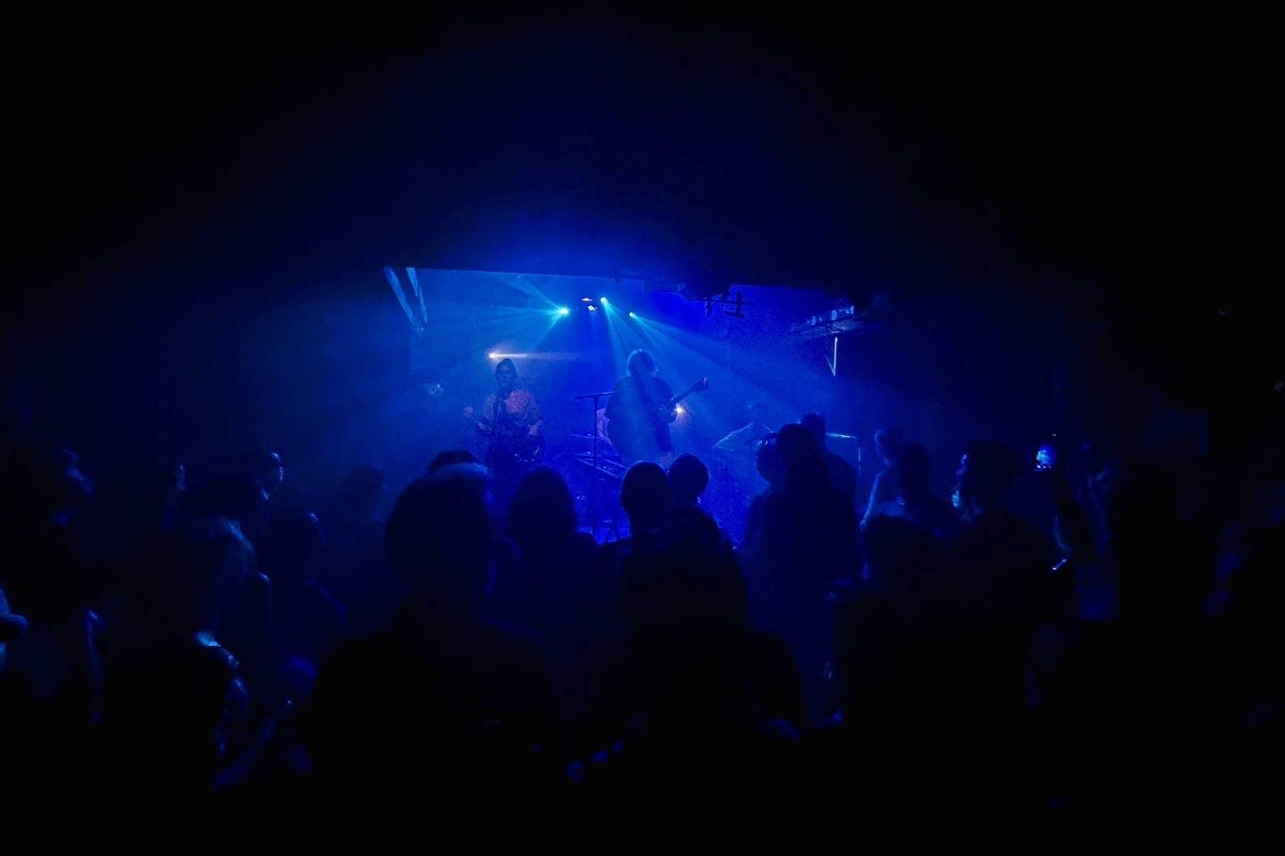 Very dark room, band playing on stage, bluish lights, but the band is barely visible, crowd in front as black shapes
