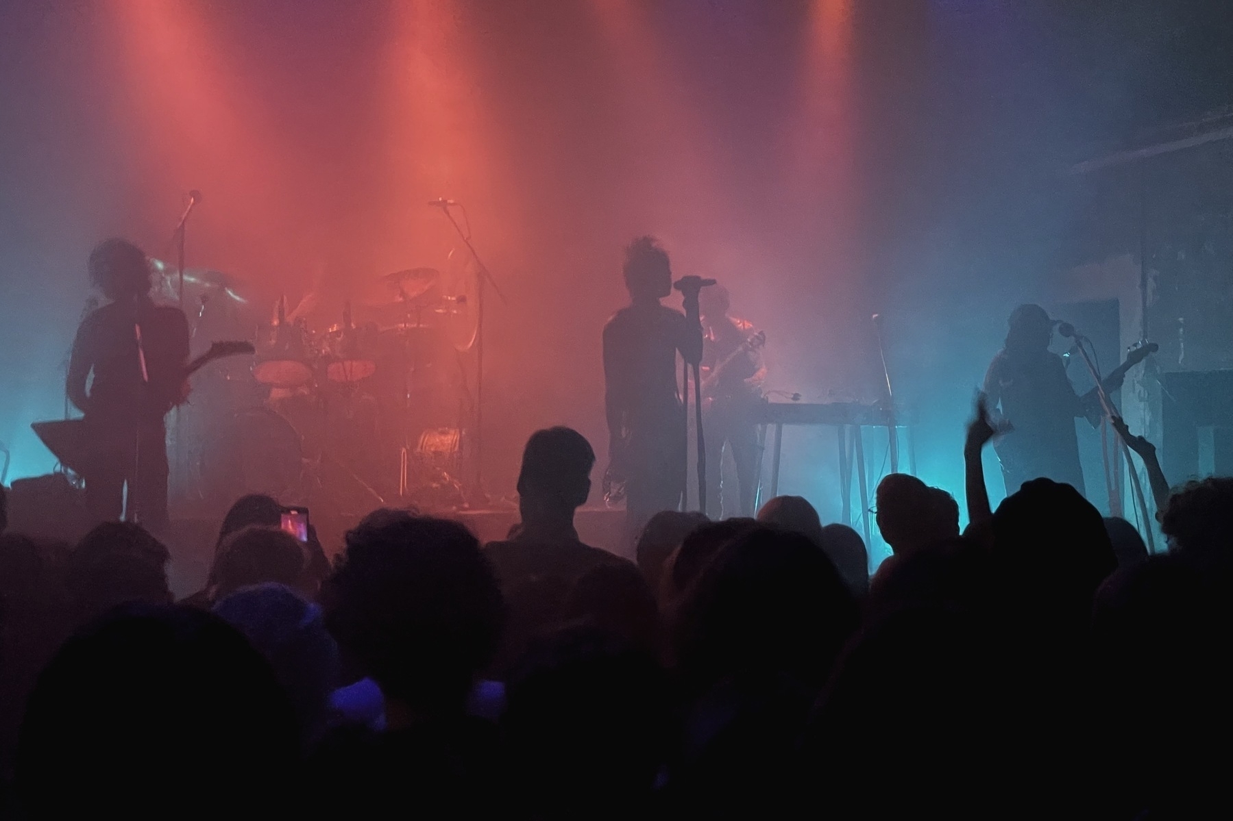 Band on stage in front of a crowd, very dark, the musicians are just shapes, the lighting is blue and red