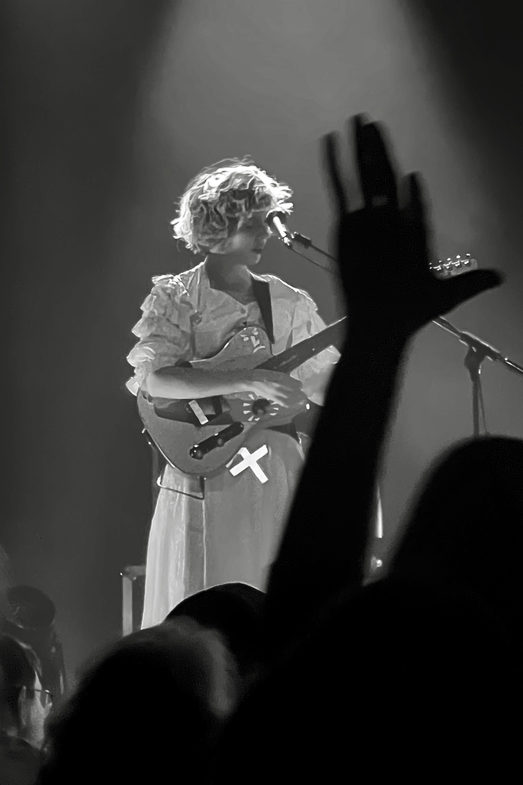 Black and white photo of a guitar player, she wears a white dress; a hand from the audience is visible blurry in the front 