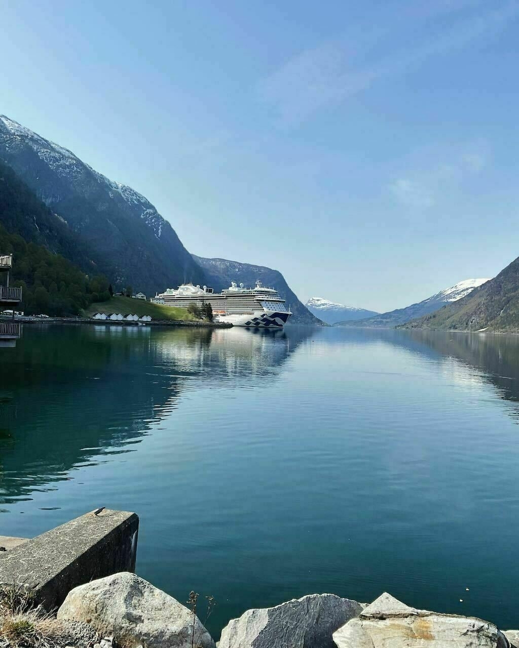 Auto-generated description: A serene fjord is depicted with a large cruise ship docked at the shoreline, surrounded by towering mountains and still waters reflecting the sky and peaks.