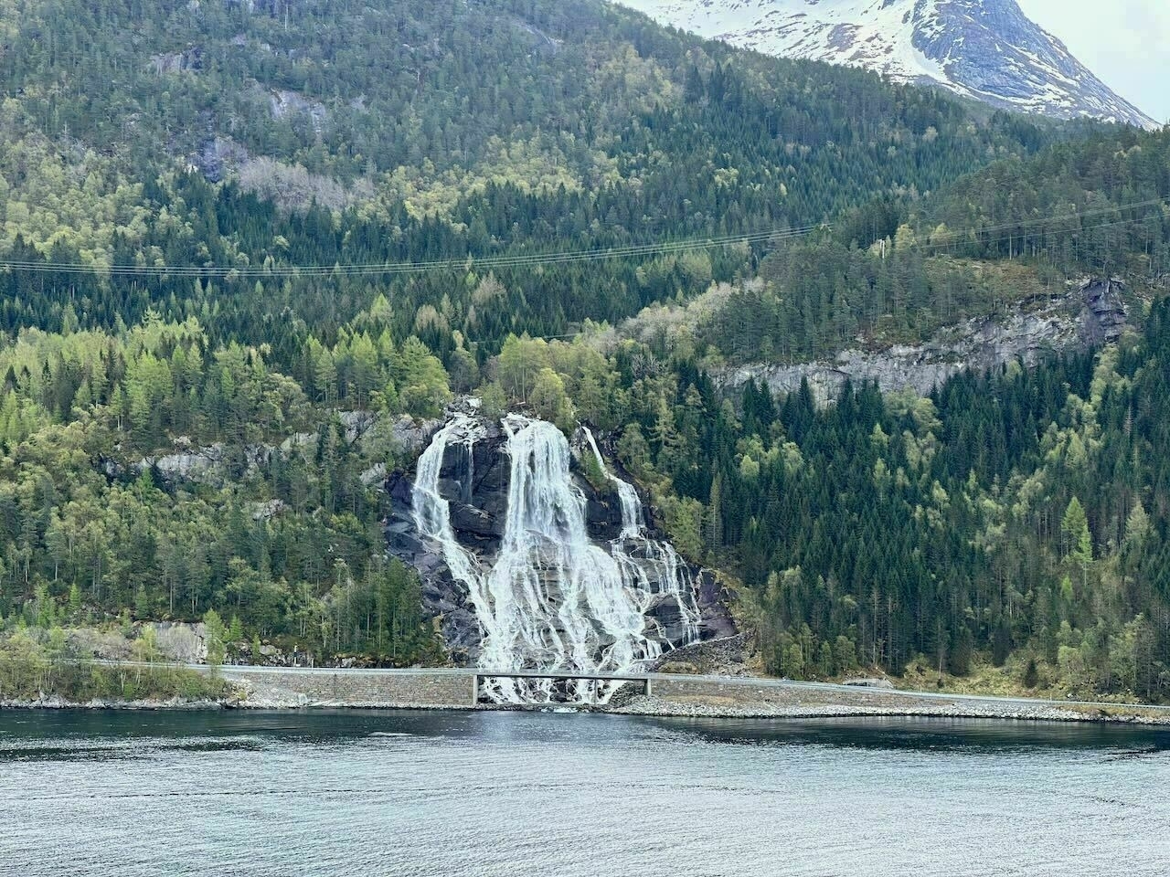 Norway waterfall cascades down a forested mountainside, framed by lush greenery and a serene body of water in the foreground.